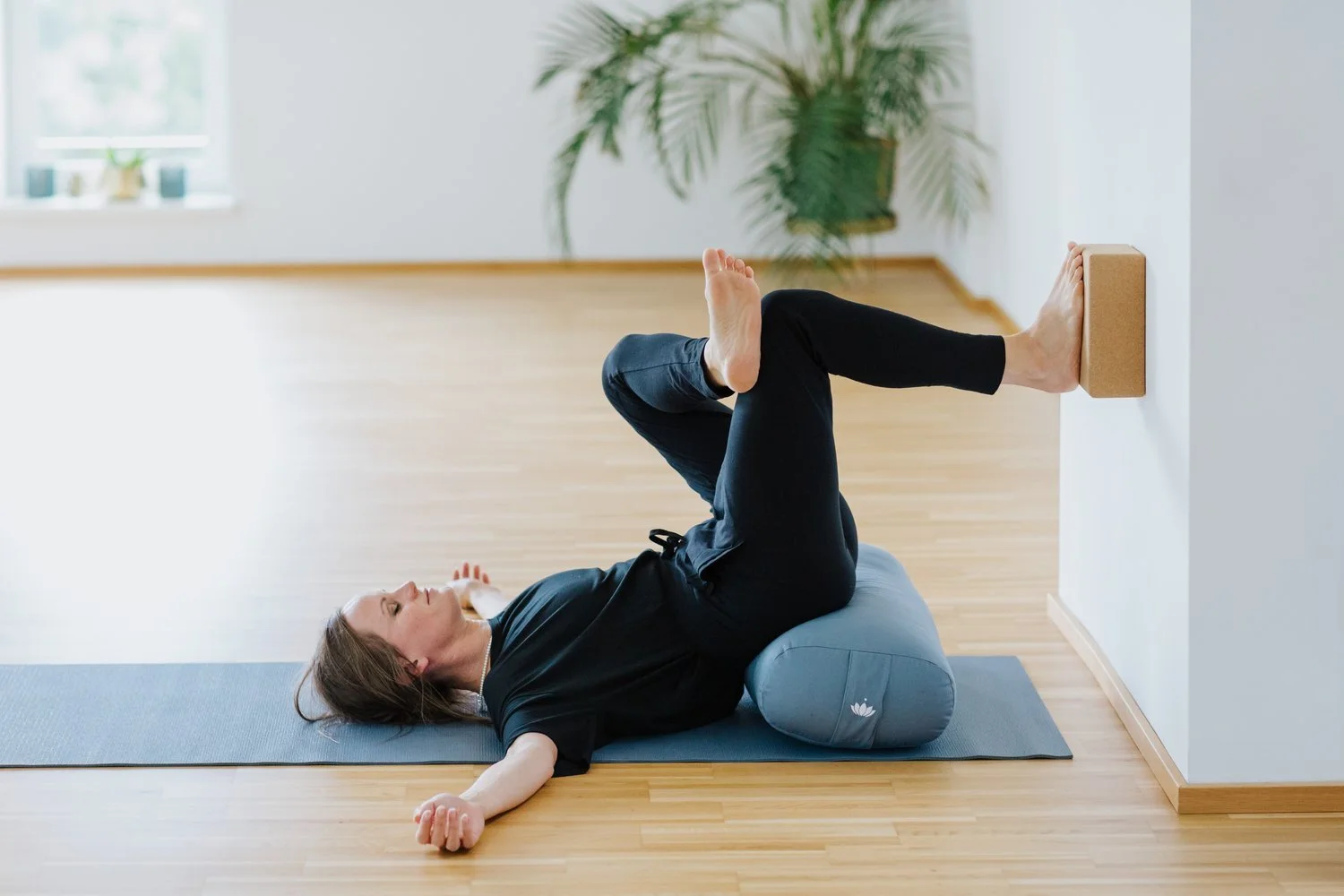 Frau liegt auf dem Rücken auf einer Yogamatte, nutzt eine Rolle unter den Knien und einen Block an der Wand zur Übung