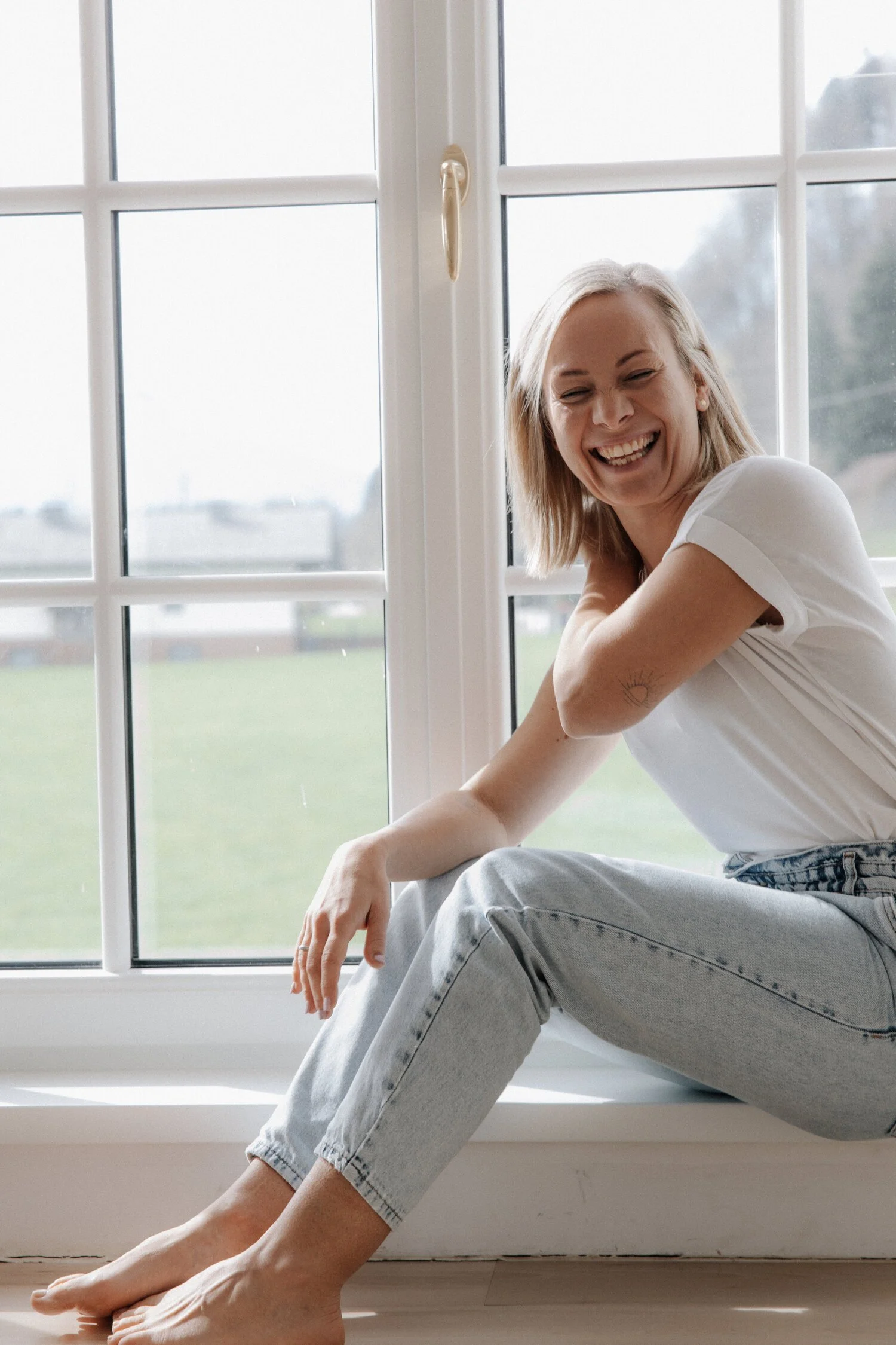 Eine lachende Frau sitzt auf einem Fensterbank, trägt einen weißen T-Shirt und helle Jeans, im Hintergrund sind große Fenster mit einer ländlichen Landschaft zu sehen.