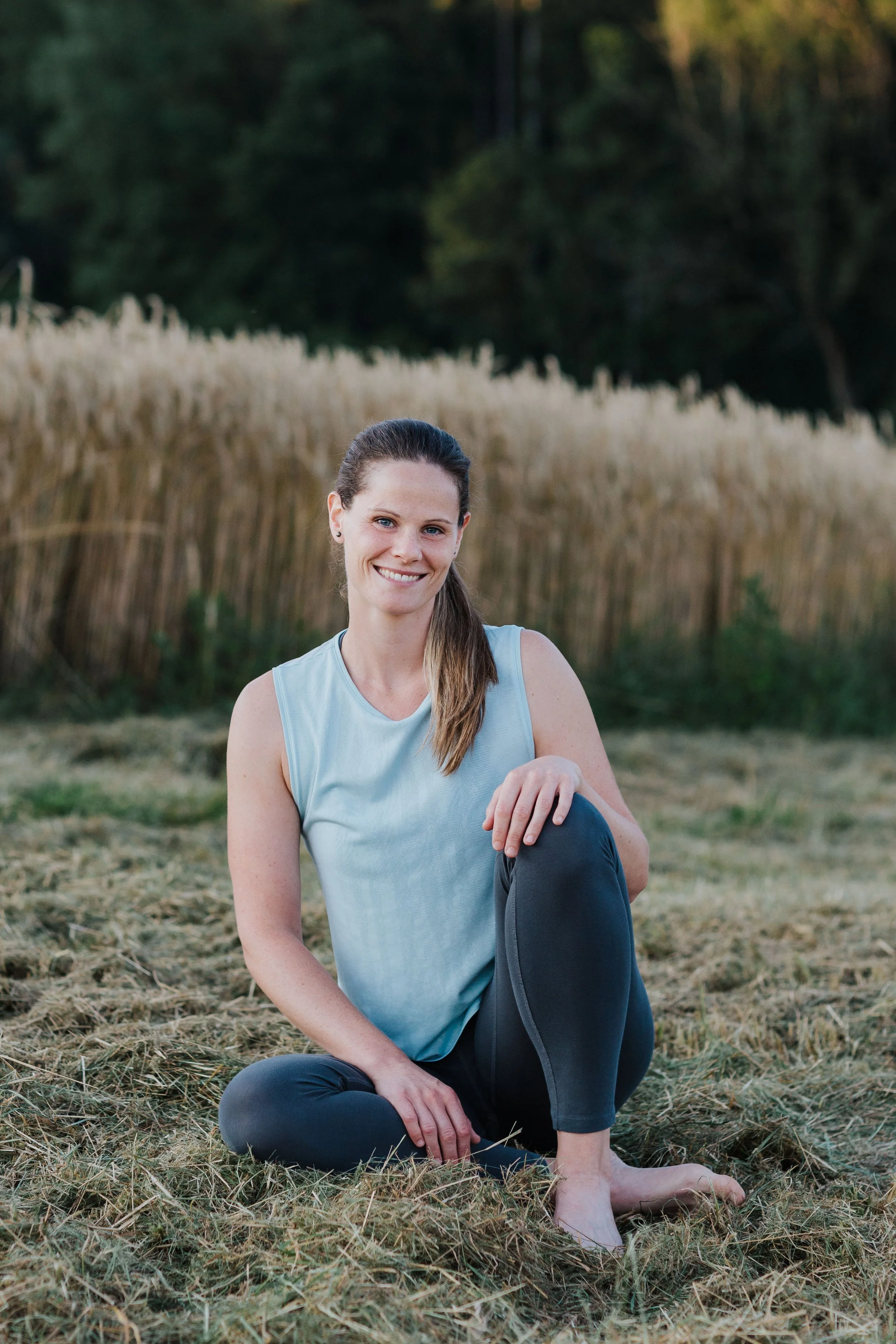 Eine lachende Frau sitzt auf dem Boden im Freien zwischen trockenem Gras, im Hintergrund Bäume und Gebüsch, trägt ein hellblaues Top und dunkle Leggings, hält ein Bein mit der Hand