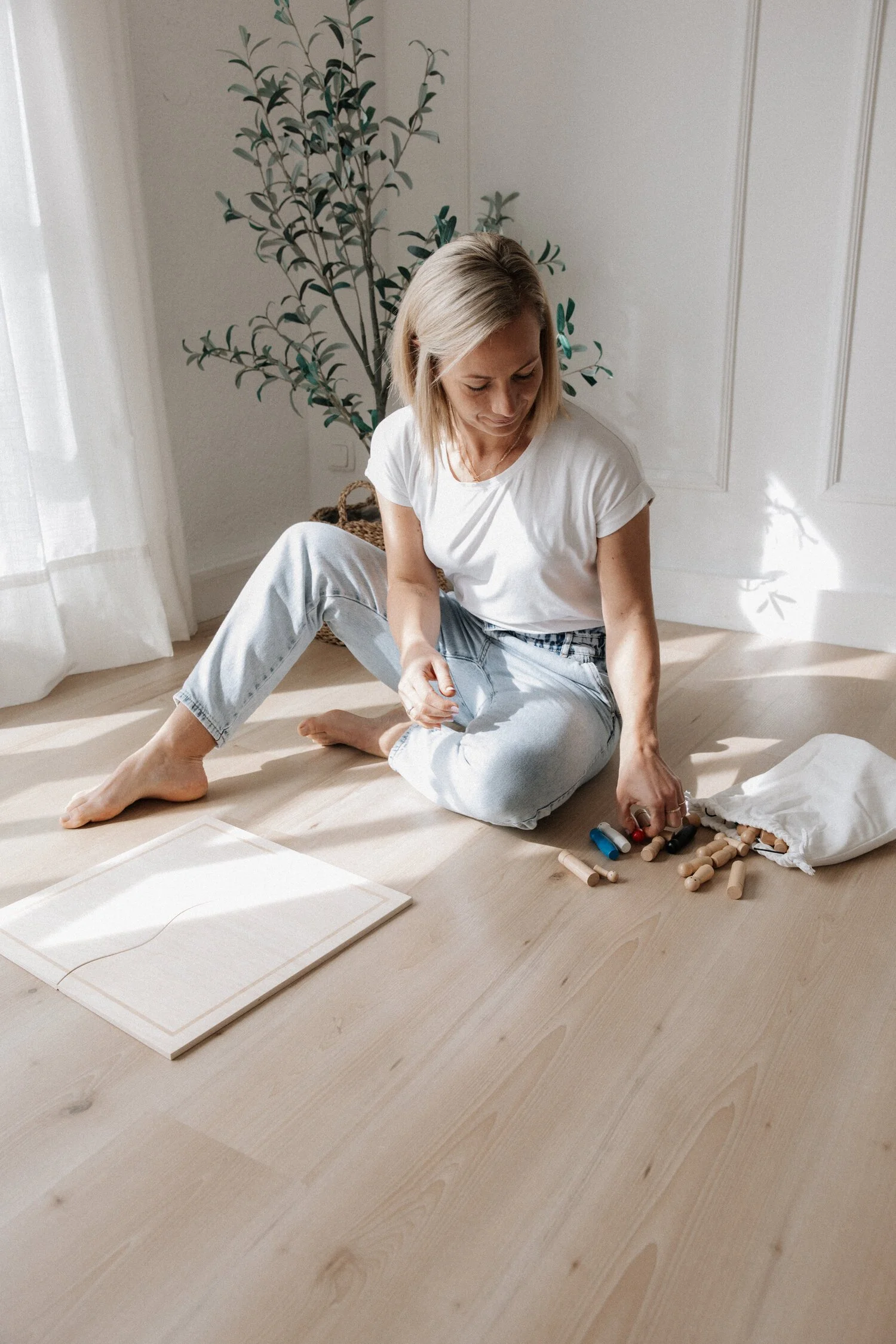 Eine Frau sitzt auf einem Holzboden und spielt mit Holzbausteinen, neben ihr liegt eine weiße Stofftasche mit weiteren Bausteinen. Im Hintergrund ist eine grüne Pflanze und weiße Vorhänge, das Sonnenlicht wirft Schatten an die Wand.