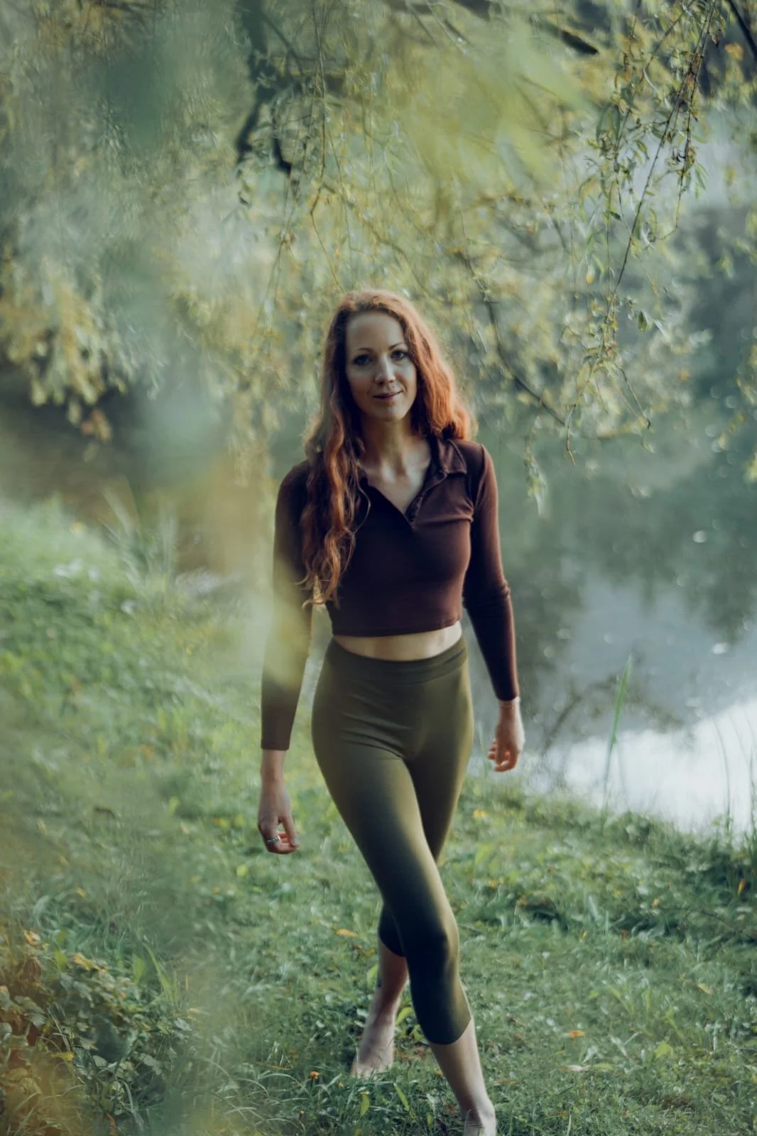 Eine junge Frau mit langen, lockigen, roten Haaren läuft barfuß durch einen See in der Natur, umgeben von grünen Bäumen und Wasser.