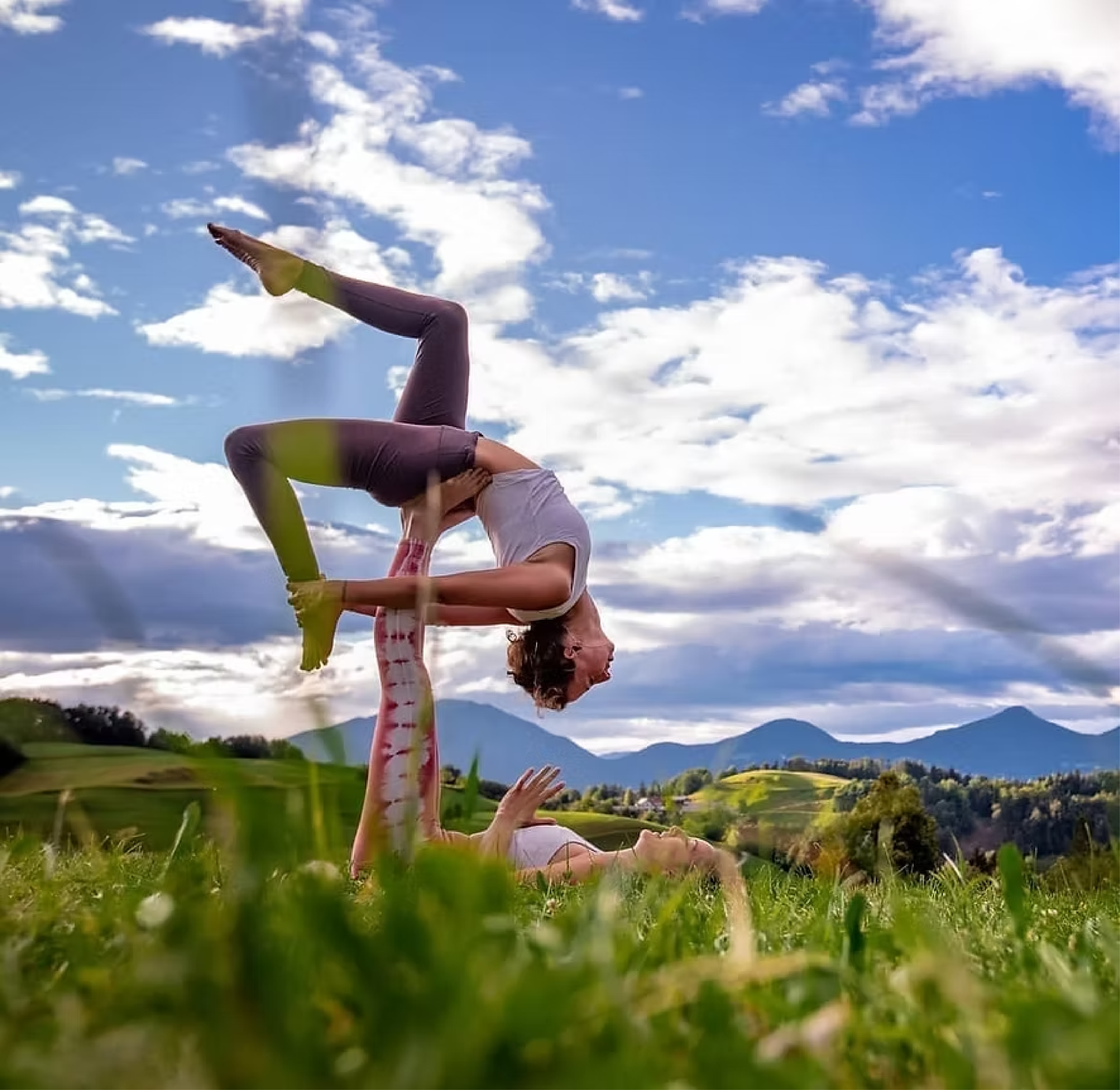 Person tut eine Partner Yoga Pose im Freien auf grünem Gras, mit blauen Himmel und Wolken im Hintergrund.