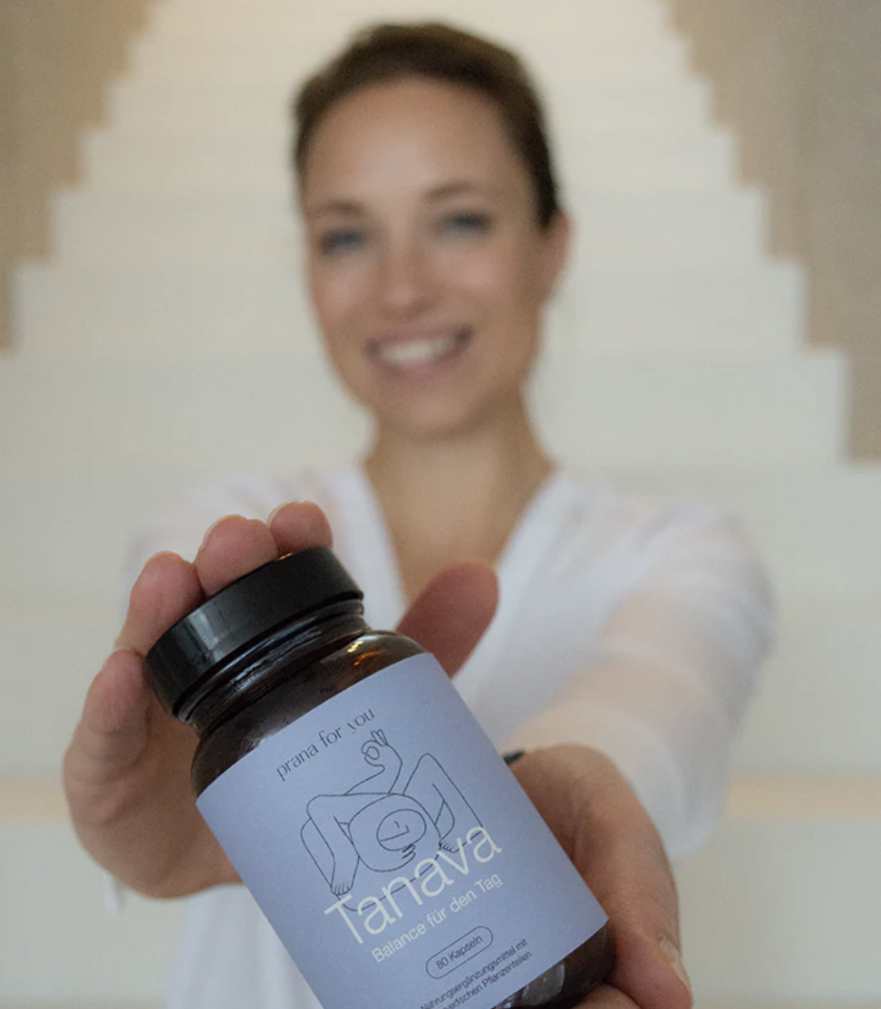 Frau hält eine Brustkapsel mit dem Label 'Tanana' und der Aufschrift 'Balance für den Tag' vor die Kamera, die im Fokus ist, während sie lächelt, im Hintergrund eine Treppe.