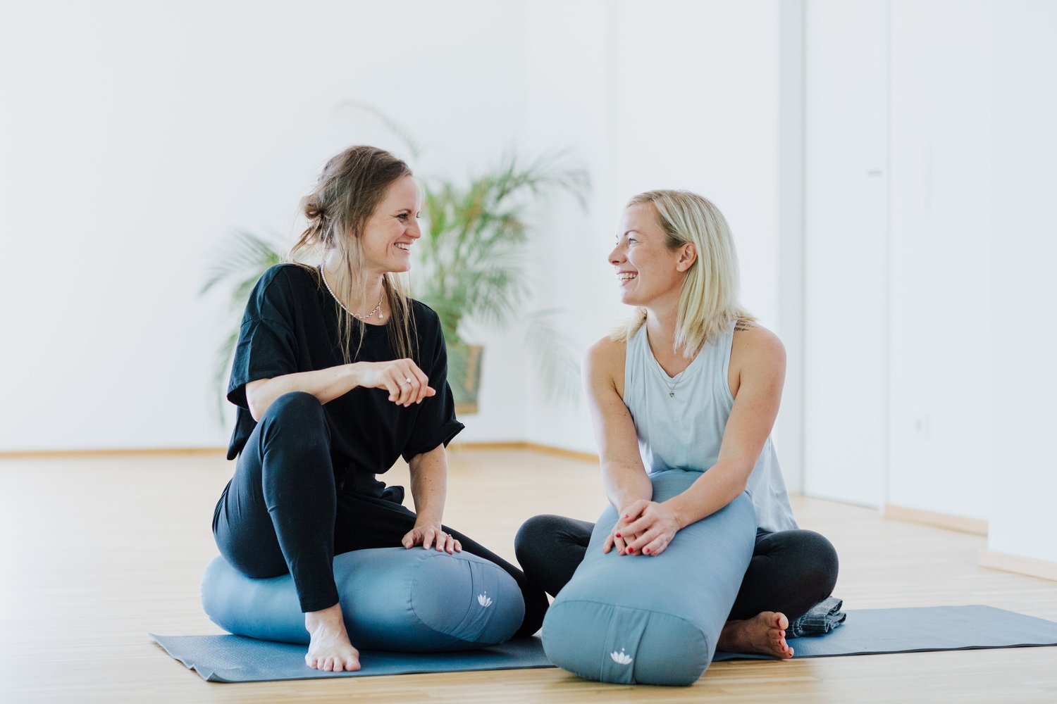 Zwei Frauen sitzen auf Yoga-Matten in einem hellen Raum, lachen und unterhalten sich.