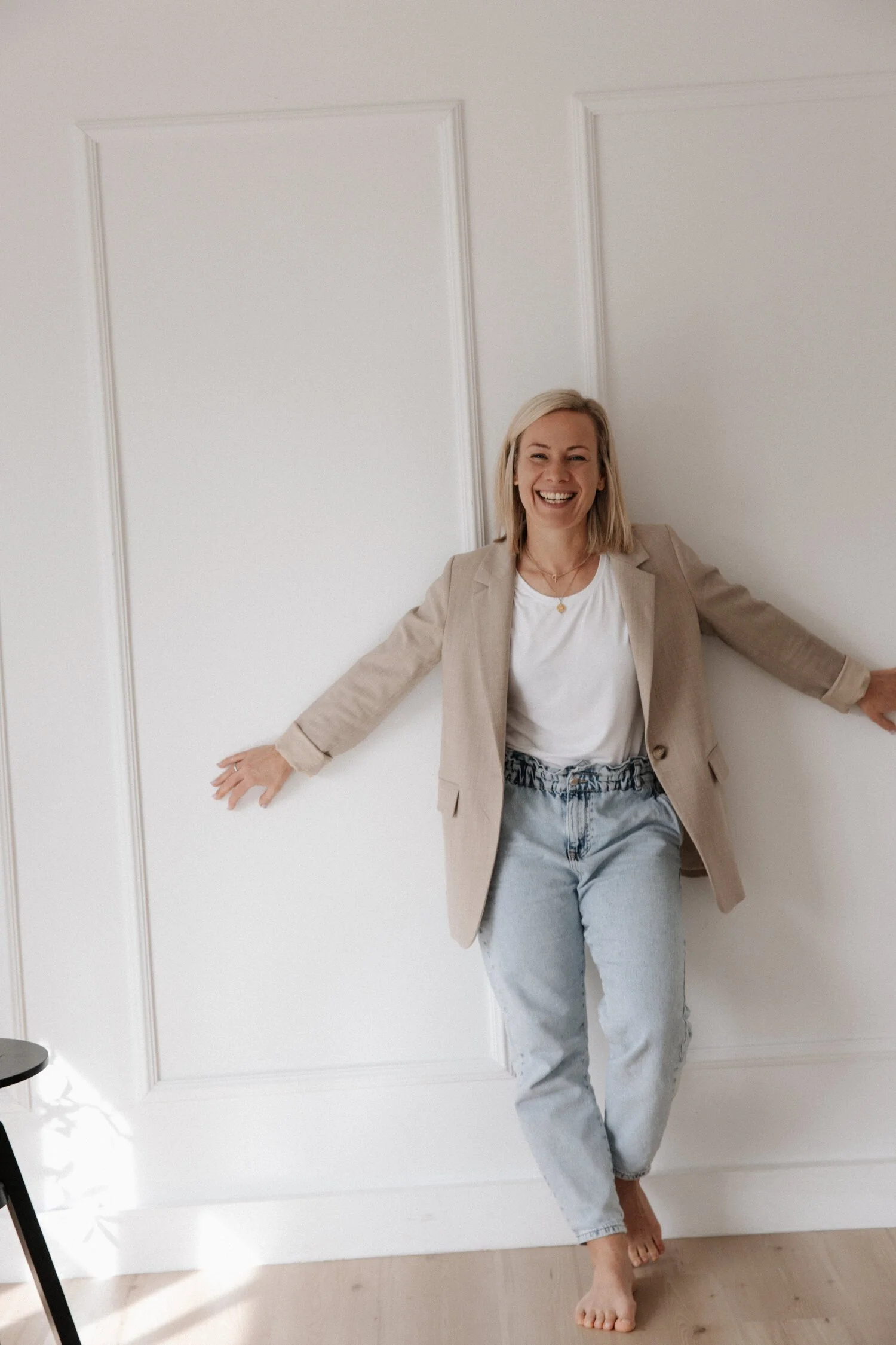 Eine lachende Frau mit blondem Haar in einem beige Hemdjacke, weißen T-Shirt und Jeanshose, die gegen eine weiße Wand lehnt, mit ausgestreckten Armen.