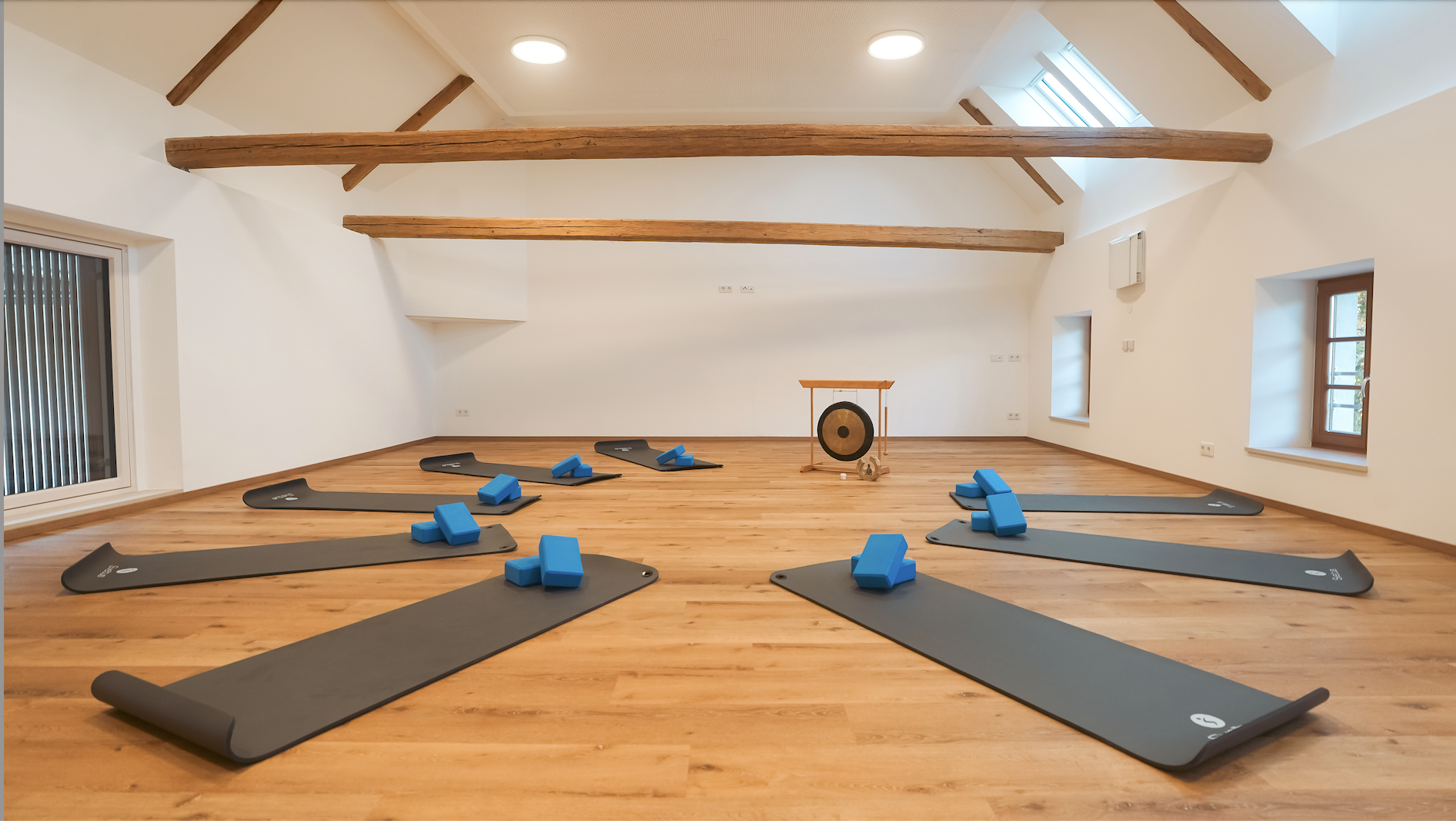 Ein Raum mit Holzboden, weißen Wänden und sichtbaren Holzbalken an der Decke. Es sind mehrere schwarze Yoga-Matten mit blauen Blöcken und dünnen Gurtbändern darauf verteilt. Im Hintergrund steht ein Gong auf einem Rahmen.