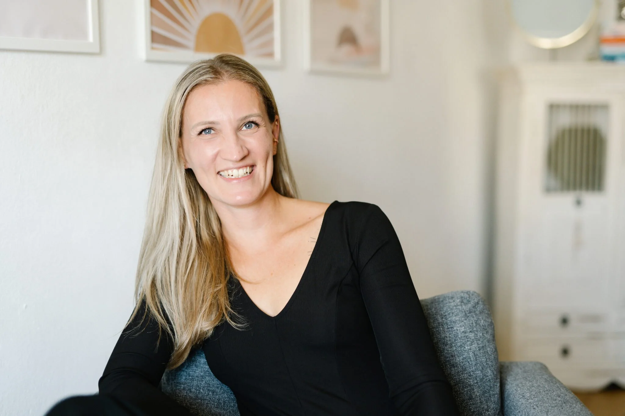 Eine lachende blonde Frau sitzt in einem gemütlichen Raum vor einer weißen Wand mit modernen Kunstwerken.