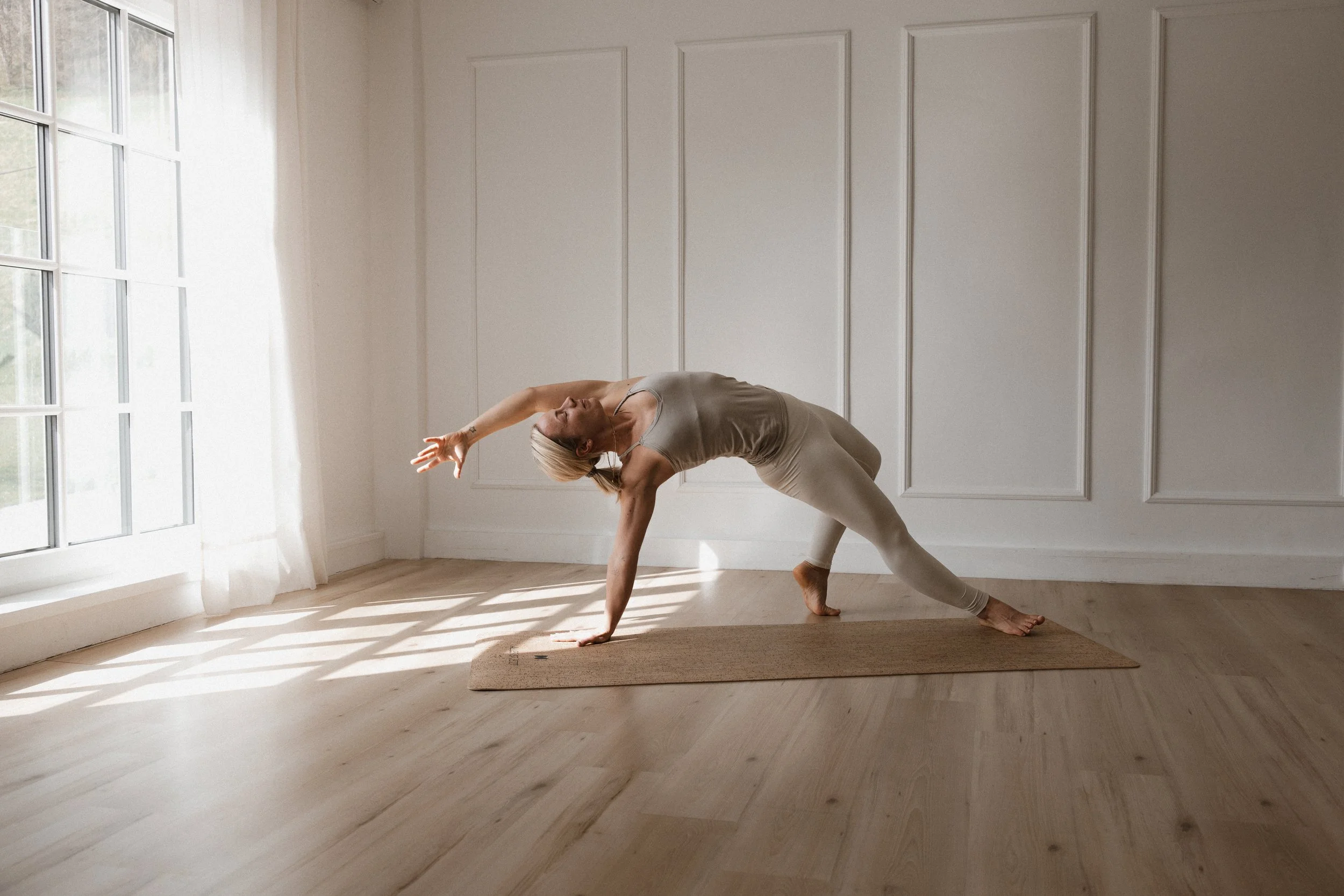 Eine Frau macht Yoga auf einer Matte vor einem großen Fenster in einem hellen Raum.