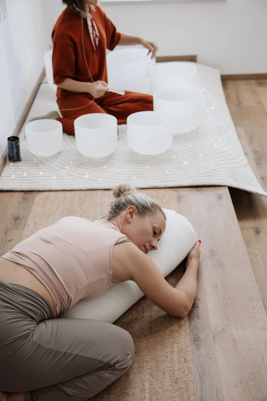 Zwei Frauen, eine sitzend mit Kristallklangschalen und die andere liegend und umarmend auf dem Boden, in einem Raum mit Holzboden und weißem Wandhintergrund.