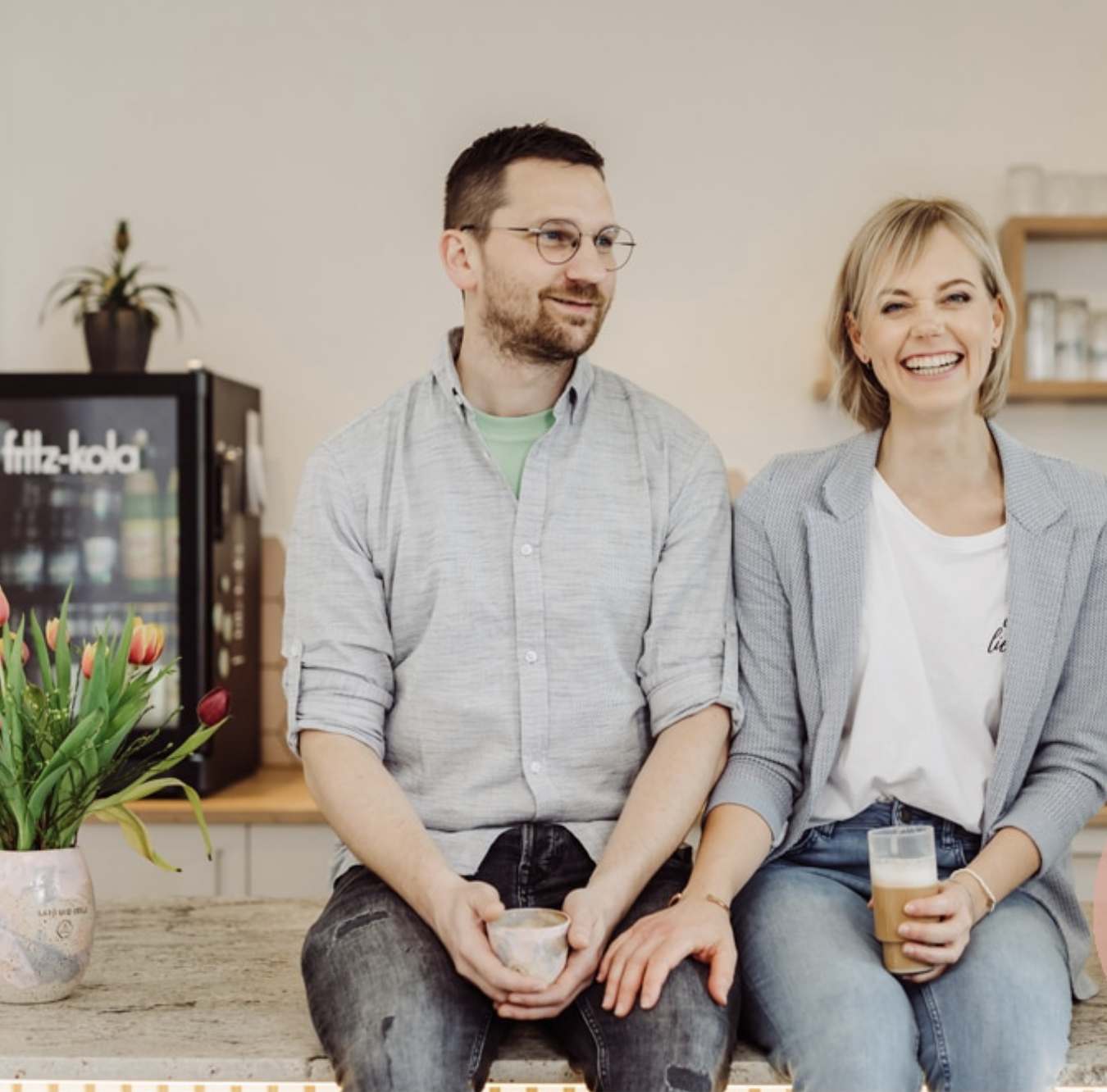 Ein Mann und eine Frau sitzen auf einer Theke in einem Café oder einer Bäckerei, die Frau hält ein Glas Kaffee, im Hintergrund eine Getränke Kühlschrank und eine Vase mit Tulpen auf dem Tisch.
