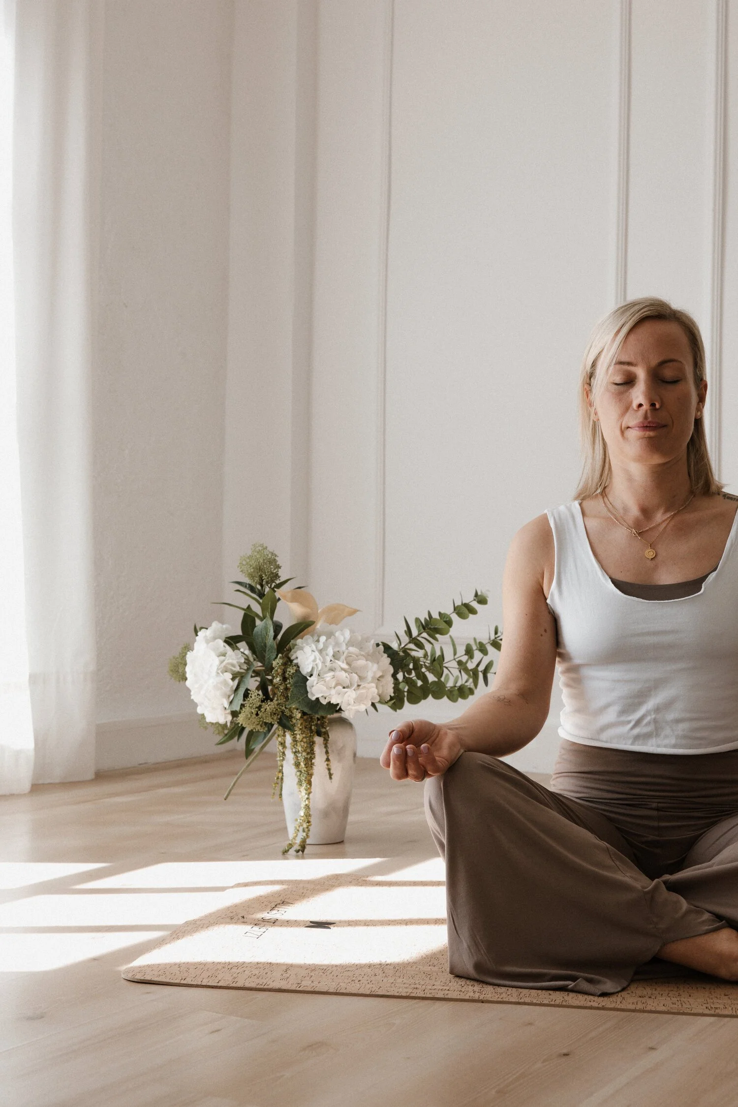 Eine Frau sitzt im Schneidersitz auf einem Yogamatte, entspannt mit geschlossenen Augen in einem hellen, minimalistischen Raum, neben einer großen Vase mit weißen Blumen.