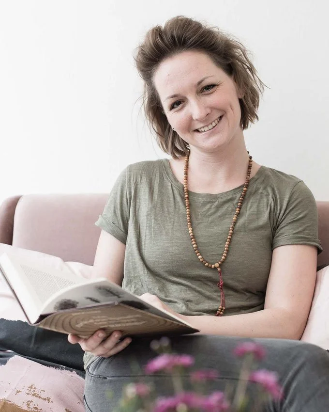 Eine junge Frau mit braunen, zerzausten Haaren, lächelnd, sitzt auf einem Sofa und liest ein Buch, trägt ein grünes T-Shirt und eine beaded Halskette.