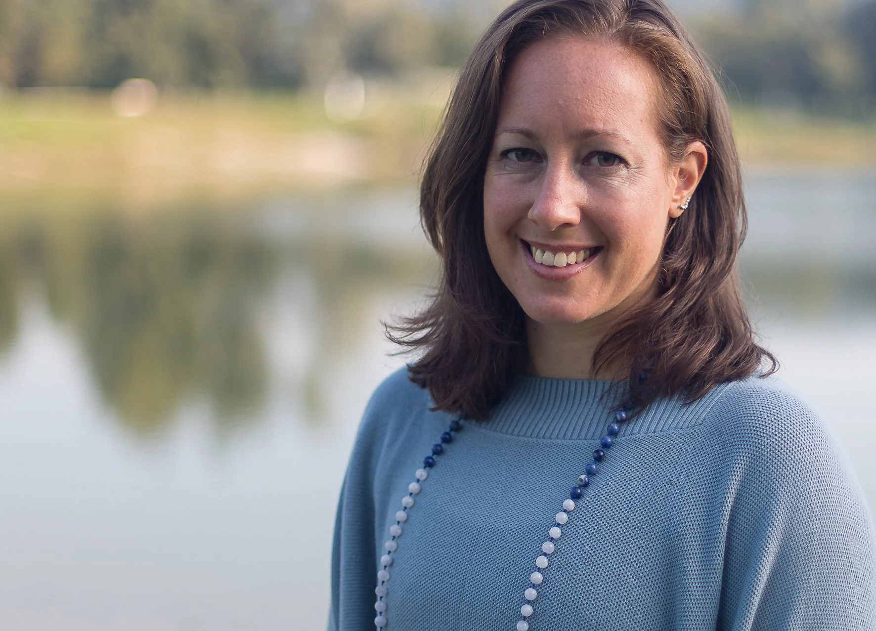Ein Portrait einer lächelnden Frau mit braunen, schulterlangen Haaren, die ein blauen Pullover und eine blau-weiße Perlenkette trägt. Sie steht vor einem ruhigen Wasser und bewaldeten Hintergrund.