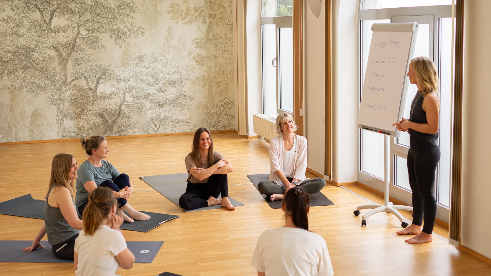 Eine Gruppe von Frauen macht Yoga im Kursraum, während die Kursleiterin vor einem Whiteboard steht.