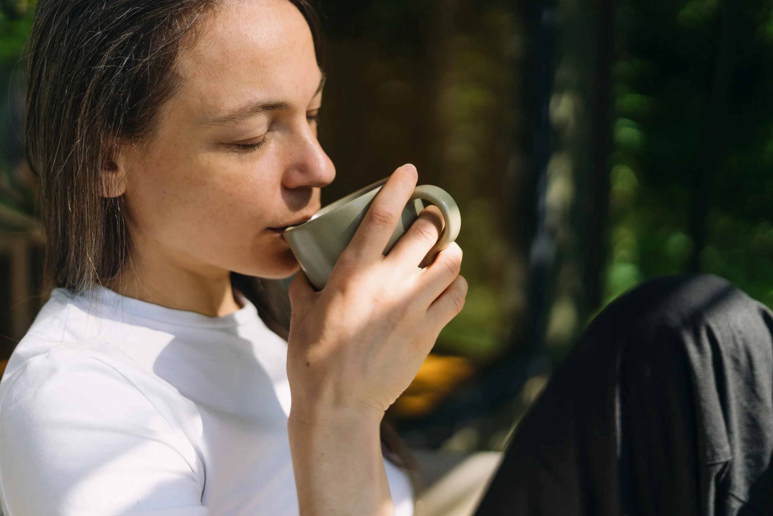 Young woman with brown hair and freckles enjoying a hot beverage outdoors on a sunny day.