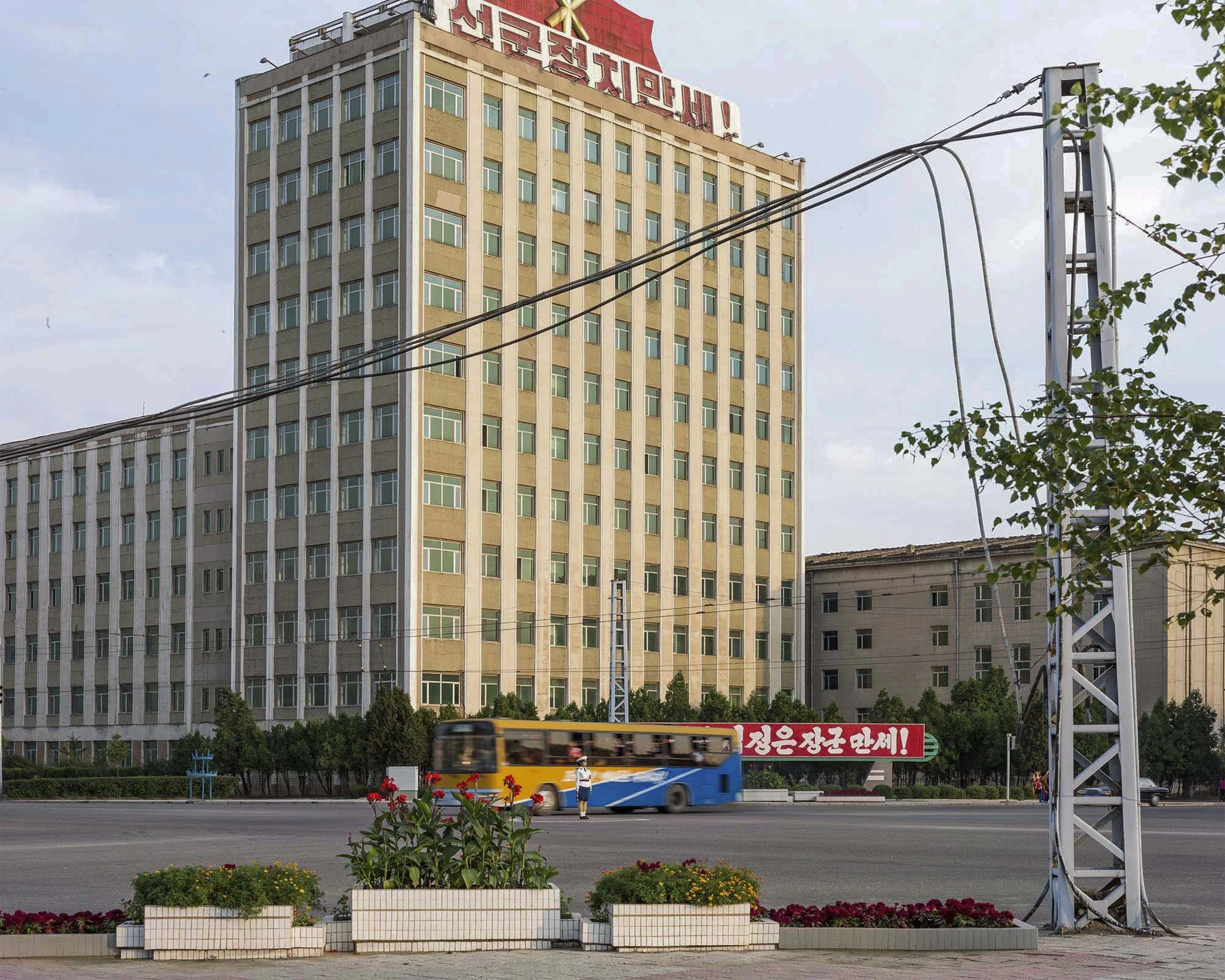 Gebäude des Zentralkomitees der Partei der Arbeit Koreas in Pjöngjang: Im Vordergrund regelt eine nordkoreanische Verkehrspolizistin in weißer Uniform den Straßenverkehr vor einem vorbeifahrenden Bus. Das repräsentative Verwaltungsgebäude der Partei 