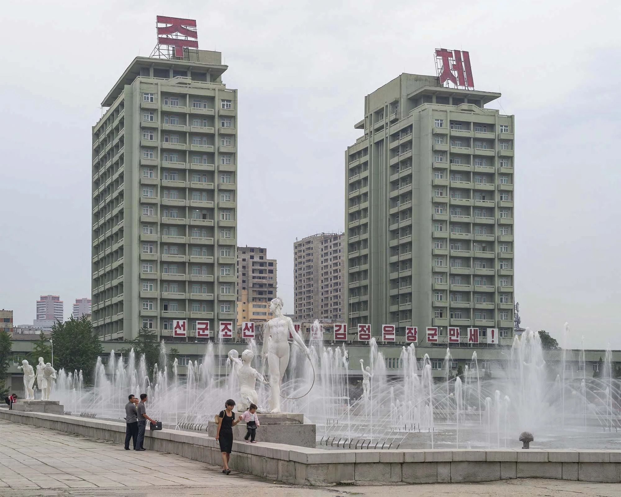Wohnkomplex und Brunnenanlage nahe der Eissporthalle in Pjöngjang, Nordkorea: Zwei charakteristische grüne Hochhäuser mit roten Dach-Slogans bilden die Kulisse für eine weitläufige Brunnenanlage mit weißen Skulpturen. Im Vordergrund flanieren Passant