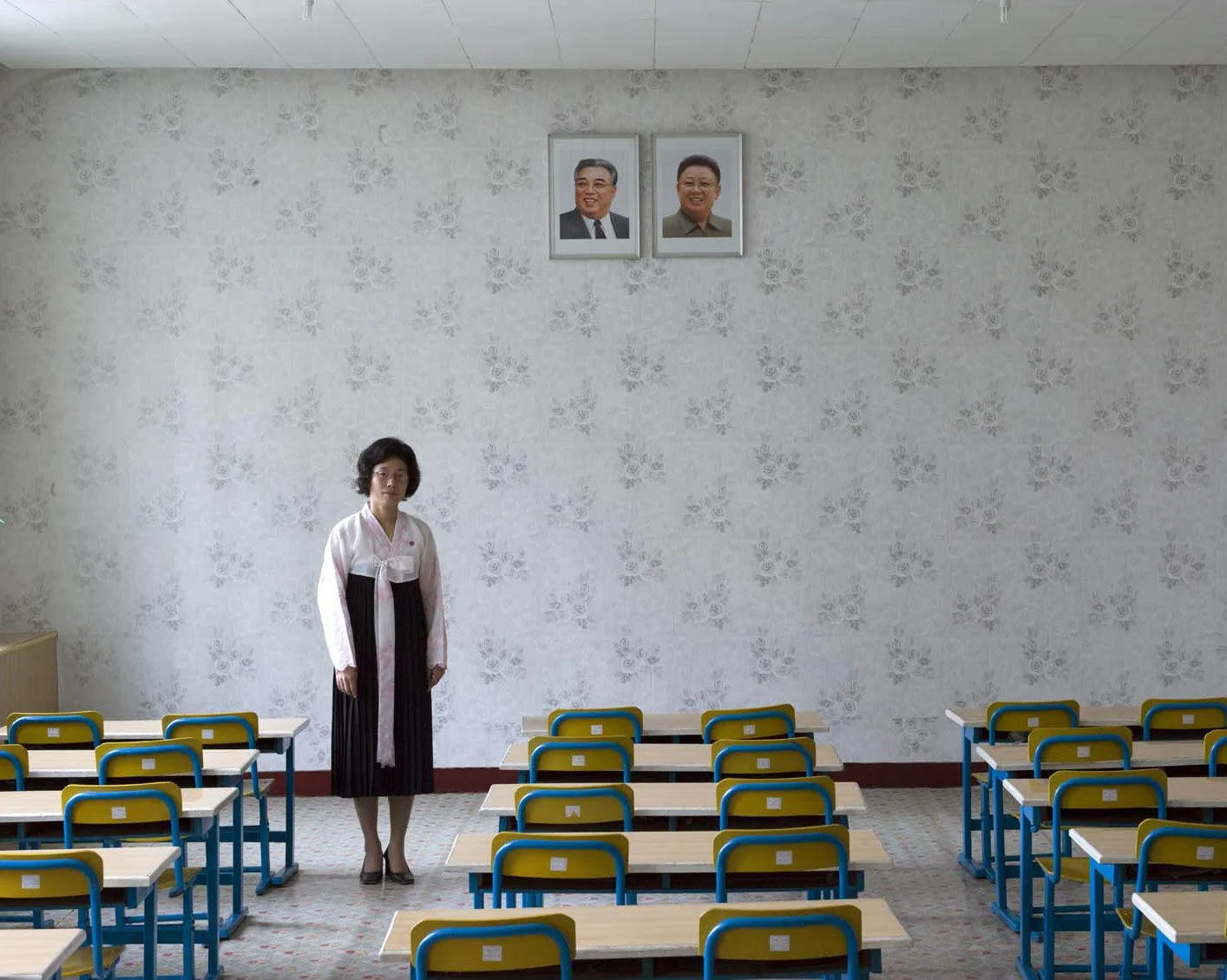 Erzieherin in einem Kindergarten in Pjöngjang, Nordkorea: Das Porträt zeigt eine Lehrerin in traditioneller Tracht (Hanbok / Chosŏn-ot) in einem leeren Klassenzimmer mit Bänken. An der gemusterten Wand hängen die Porträts der Führungspersönlichkeiten