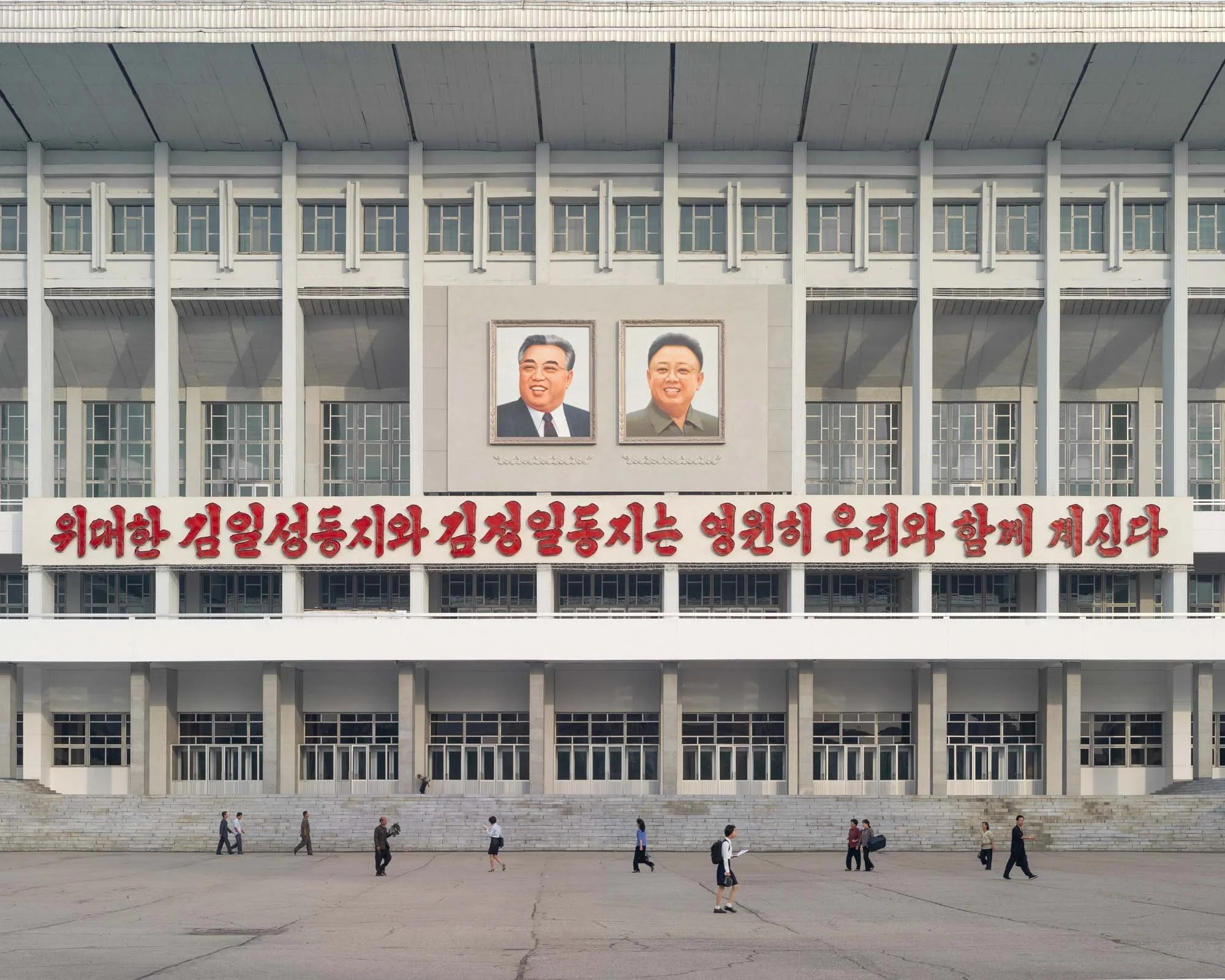 Pyongyang Indoor Stadium (Sporthalle Pjöngjang), Nordkorea: Die imposante Fassade der großen Sporthalle nahe der Eissporthalle zeigt monumentale Porträts von Kim Il-sung und Kim Jong-il sowie einen roten Propagandaslogan. Vor dem Gebäude kreuzen einz
