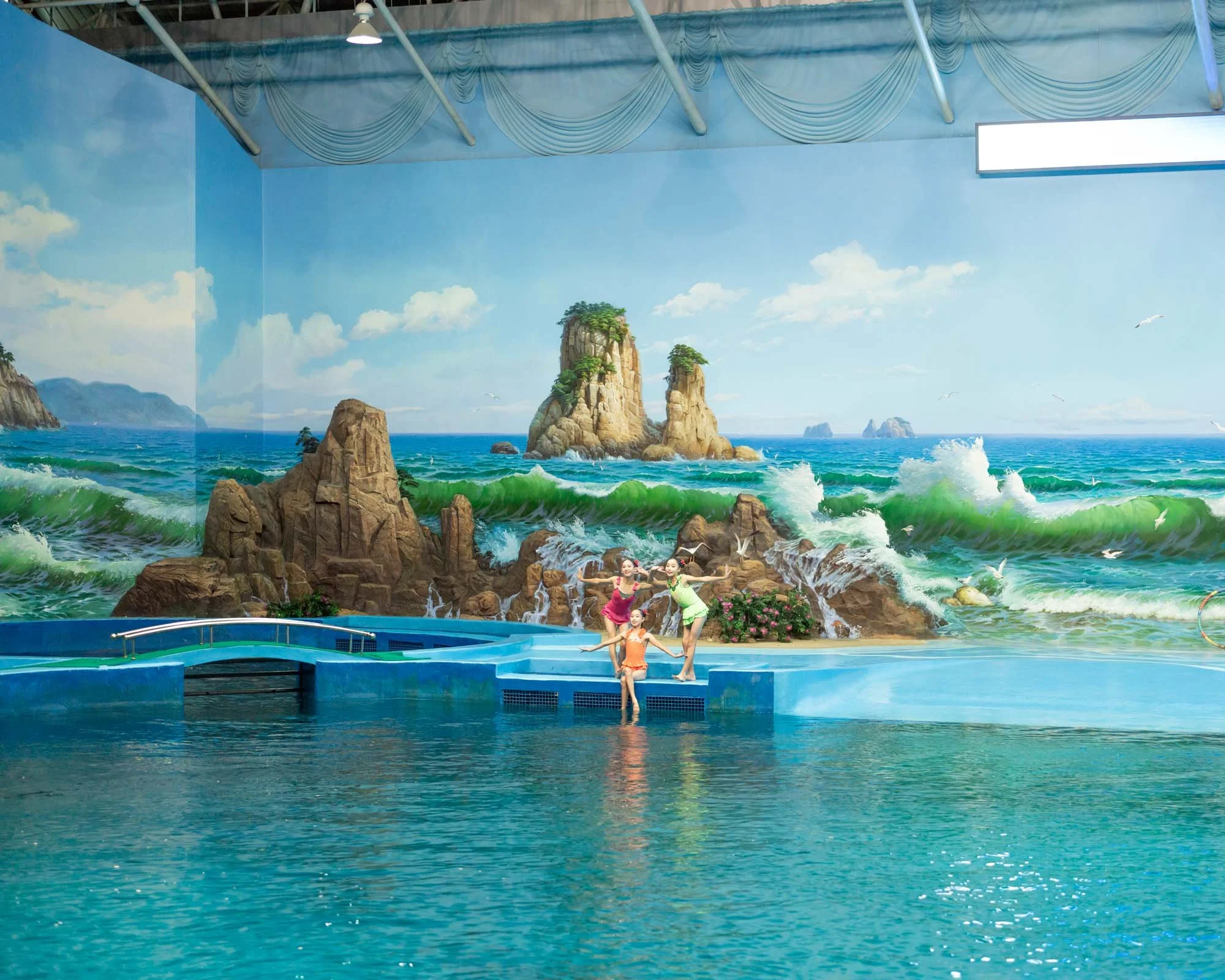Hallenbad im im Delphinarium)in Pjöngjang, Nordkorea: Drei junge Schwimmerinnen posieren am Beckenrand vor einer fotorealistisch gemalten Kulisse einer kilometerweiten Küstenlandschaft mit Meeresfelsen und Wellen. Die aufwendige Illusionsmalerei verl