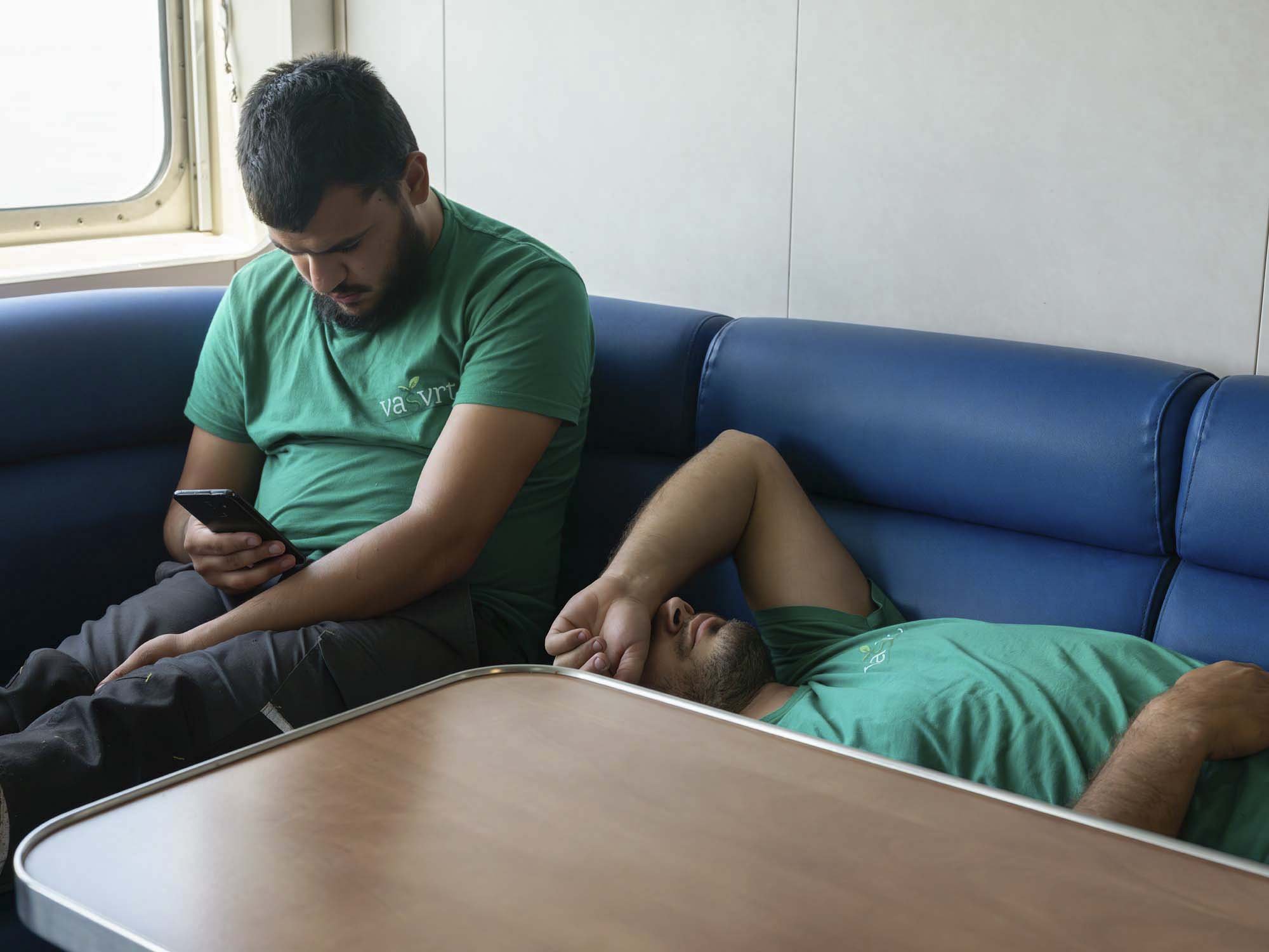 Zwei Männer, einer sitzt, der andere liegt schlafend auf einem Sitz im Krankenzimmer. Der sitzende Mann schaut auf sein Smartphone, beide tragen grüne T-Shirts.