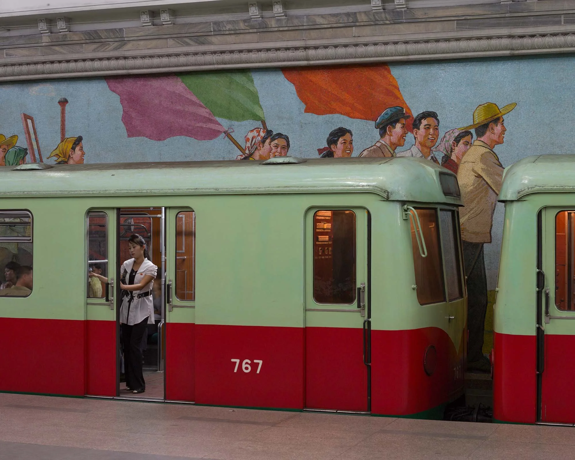 Historische Berliner U-Bahn-Waggons in der Metro Pjöngjang, Nordkorea: Ein alt-grün und rot lackierter Zug der ehemaligen Berliner Verkehrsbetriebe (Gisela / Baureihe G) steht an einem Bahnsteig mit einem farbenfrohen Mosaik, das Arbeiter und Flaggen