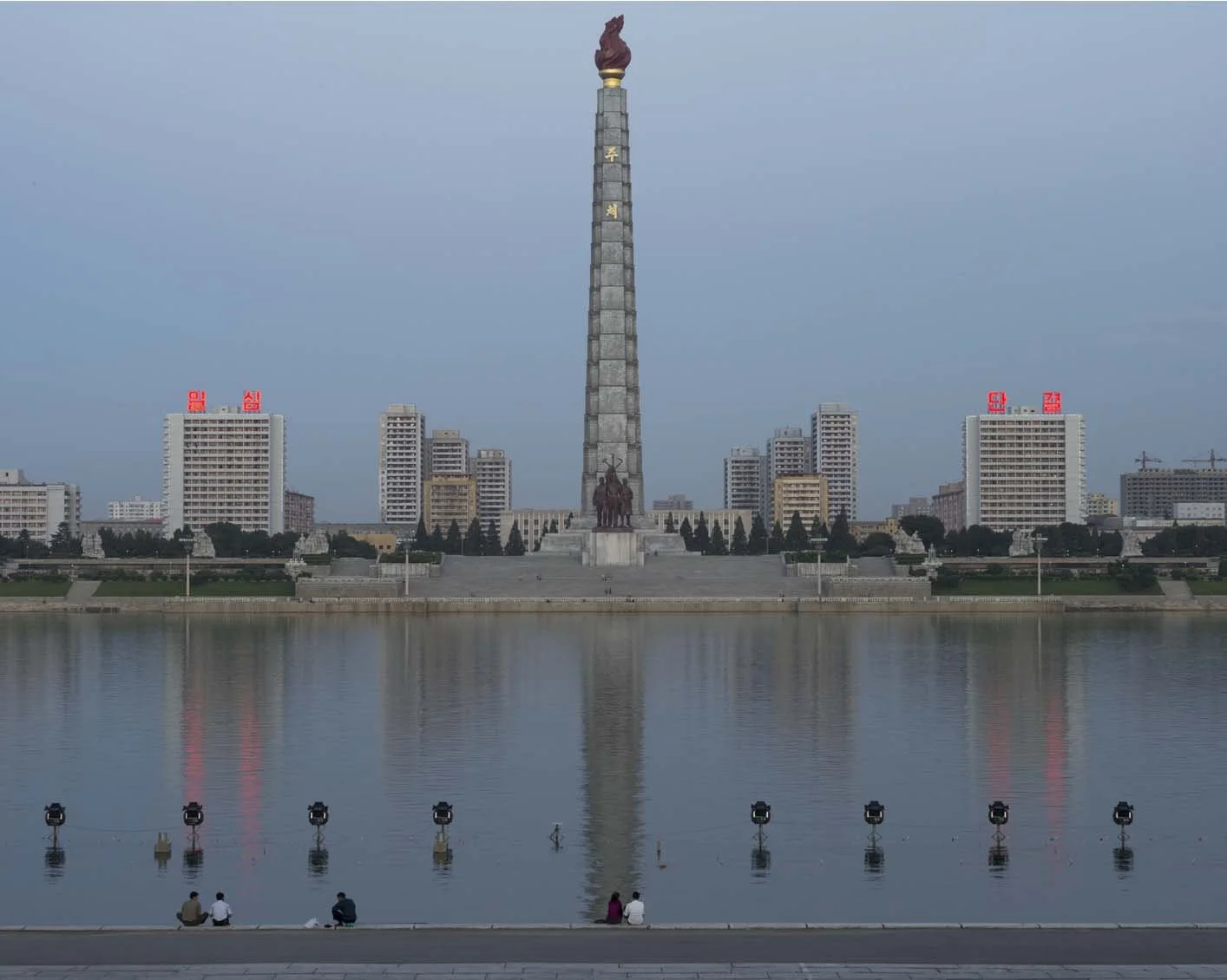 Blick über den Taedong-Fluss auf den Juche-Turm in Pjöngjang, Nordkorea: Symmetrische Ansicht des 170 Meter hohen Monuments der Juche-Ideologie von der Uferpromenade aus. Das Bauwerk spiegelt sich im stillen Wasser des Flusses, während im Vordergrund