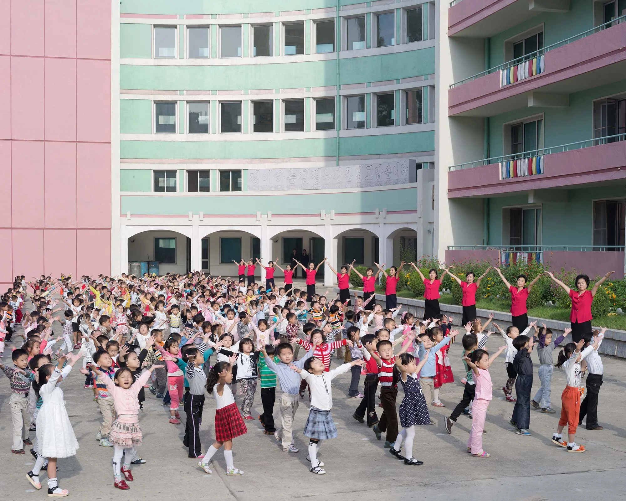 Morgengymnastik im Innenhof eines Kindergartens in Pjöngjang, Nordkorea: Hunderte Kinder machen synchron Bewegungsübungen unter der Anleitung ihrer Erzieherinnen im gepflasterten Außenbereich. Das moderne, in Pastellfarben gehaltene Gebäude trägt am 