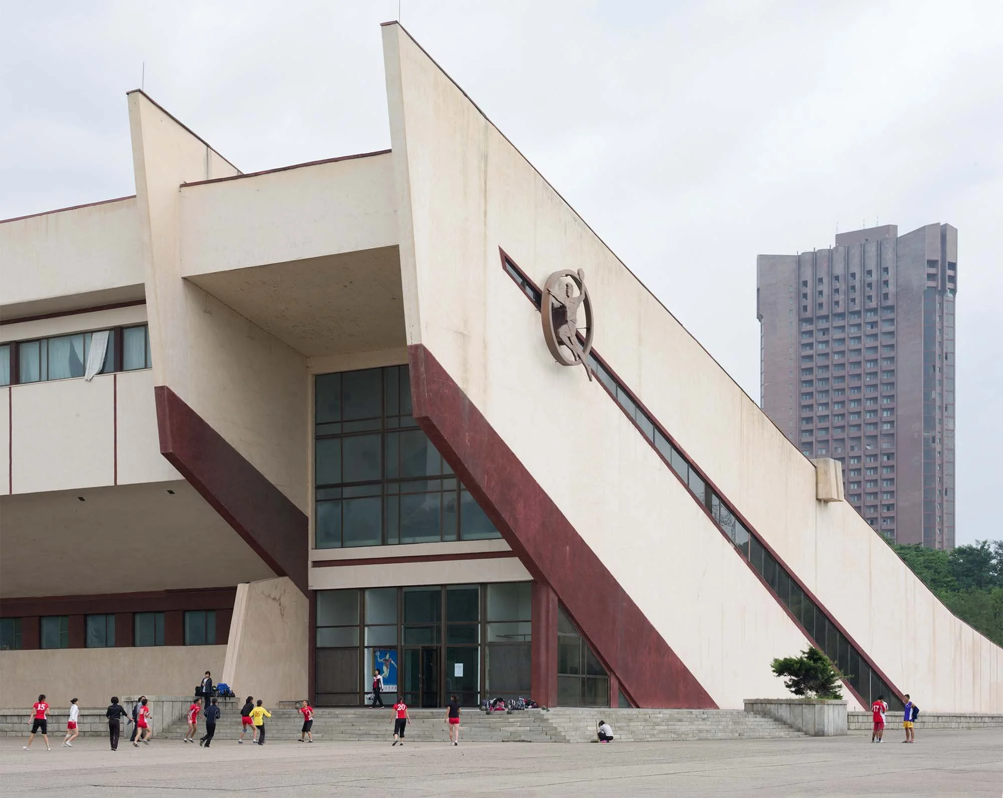 Sporthalle der Chongchun-Sportstraße in Pjöngjang, Nordkorea: Das Gebäude mit dynamisch ansteigender keilförmiger Architektur gehört zum Sportkomplex in der Chongchun-Straße. Vor dem Eingang trainieren Jugendliche auf dem Vorplatz, während im Hinterg
