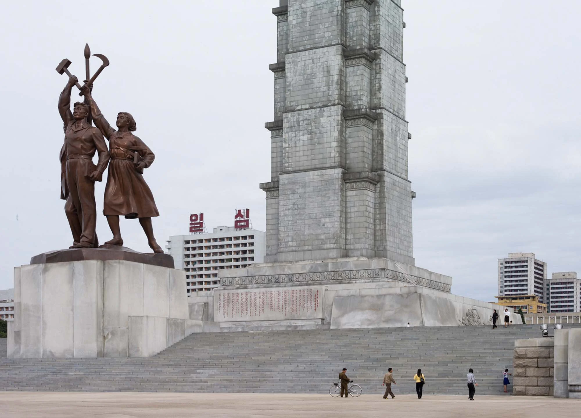 Sockel und Drei-Figuren-Statue des Juche-Turms in Pjöngjang, Nordkorea: Nahaufnahme der Basis des 170 Meter hohen Granitturms. Links erhebt sich die charakteristische 30 Meter hohe Bronzestatue, die drei Arbeiter mit emporgehobener Hammer, Sichel und