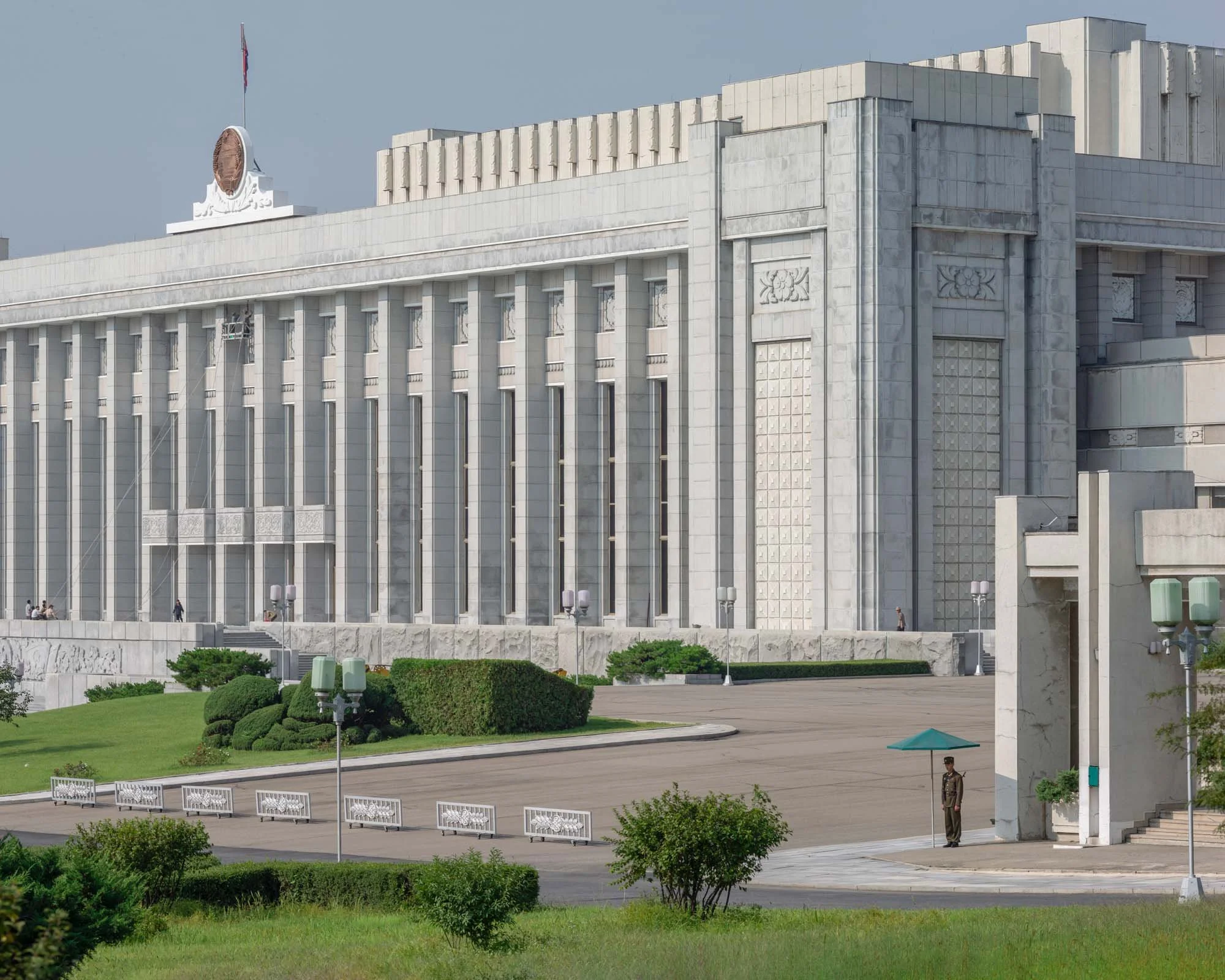 Mansudae-Kongresshalle (Parlament) in Pjöngjang, Nordkorea: Der imposante Neoklassizismus-Bau dient als Tagungsort der Obersten Volksversammlung. Die weiße Granitfassade mit ihren massiven Säulen dominiert den Platz, während im Vordergrund ein Wachpo