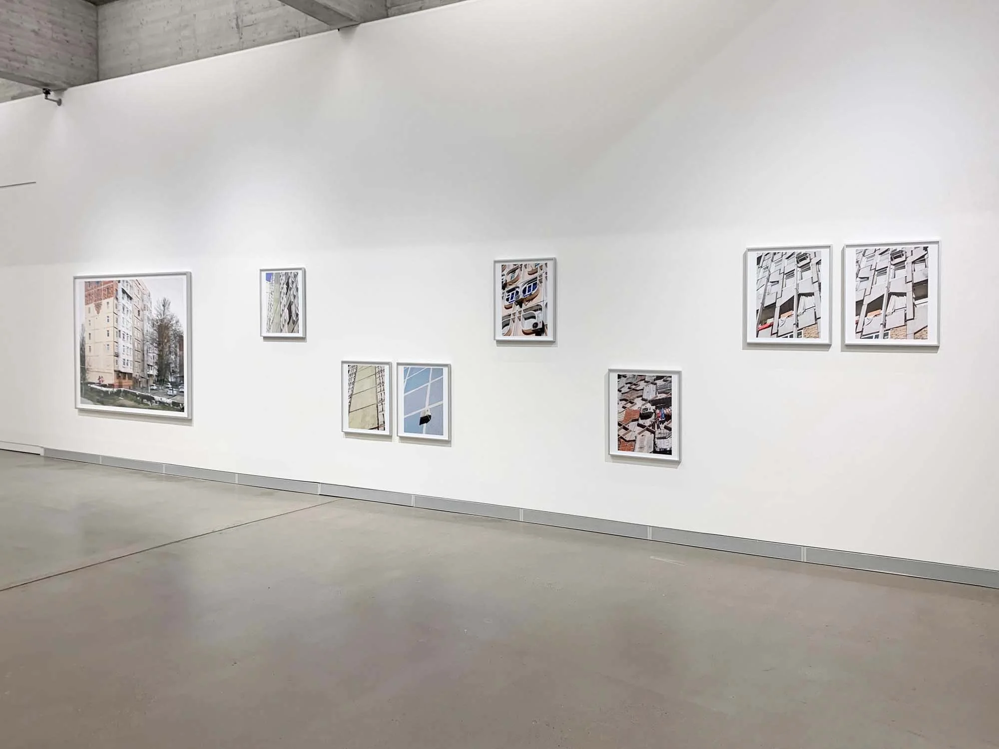 Fotografie einer Kunstgalerie mit weißen Wänden, auf denen mehrere moderne Stadt- und Architekturaufnahmen in Rahmen hängen.
