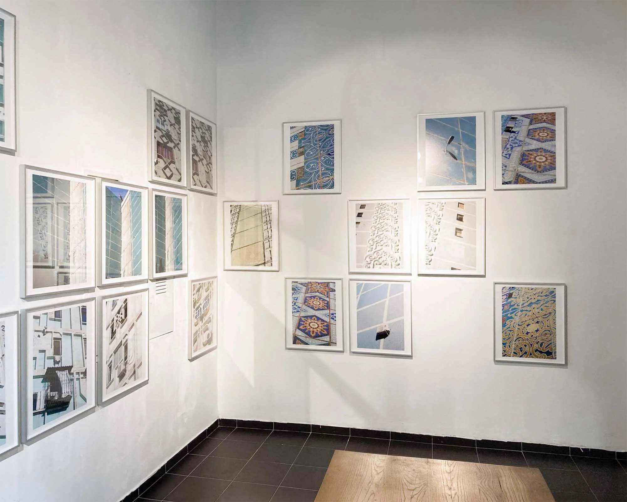 Stillleben einer Kunstgalerie mit weißen Wänden, an denen mehrere Rahmen mit Fotografien und Kunstwerken hängen. Der Boden ist dunkel gefliest, in einer Ecke ein kleiner Bereich mit Holzboden. Ein Sitzmöbel ist teilweise sichtbar.
