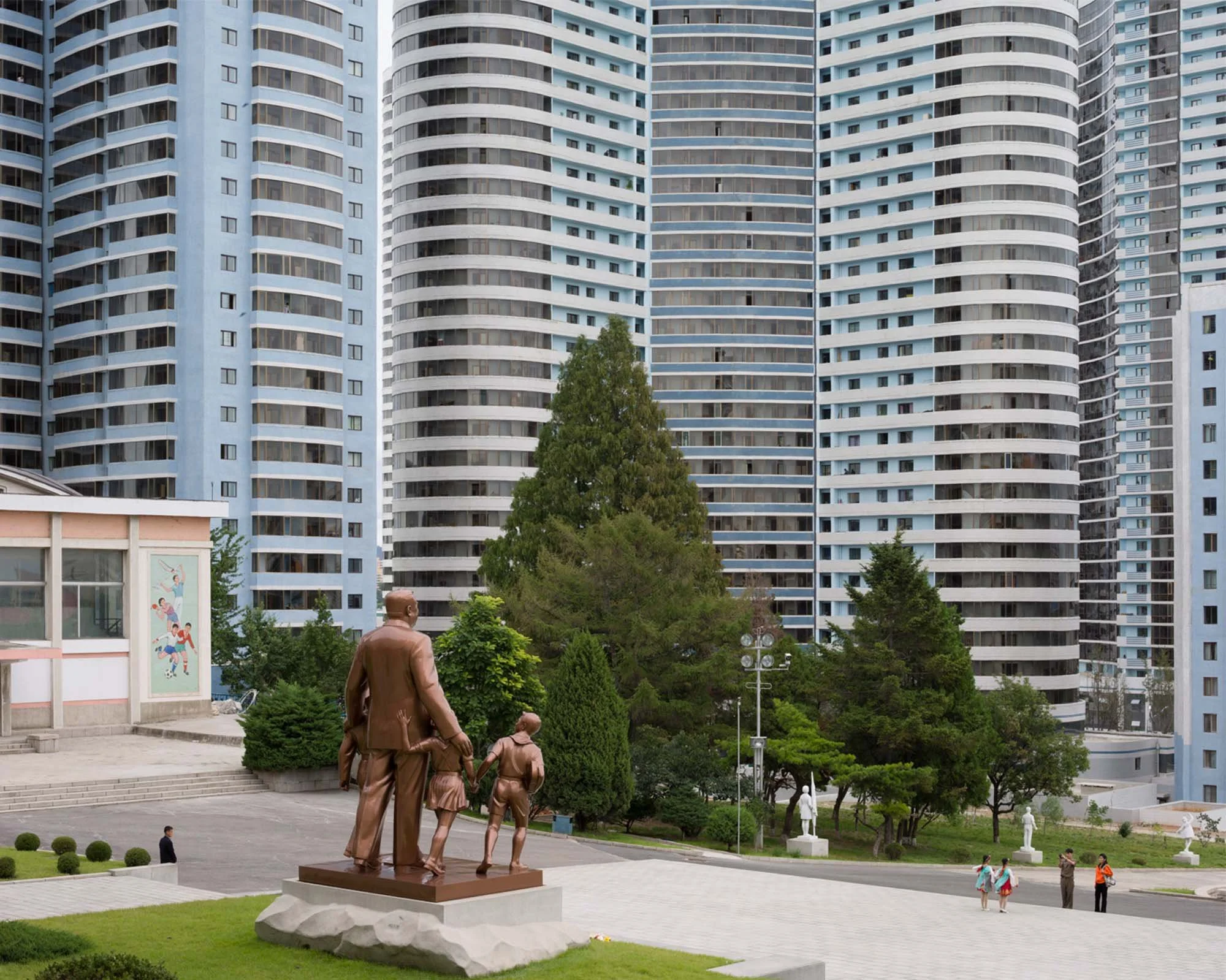Statue von Kim Il-sung mit Kindern nahe dem alten Schülerpalast (Jangchang) in Pjöngjang, Nordkorea: Die Bronze-Statue zeigt den Staatsgründer im Kreis von Kindern, aufgestellt vor der kulissenartigen Modernisierung der Changjon-Straße mit ihren gesc