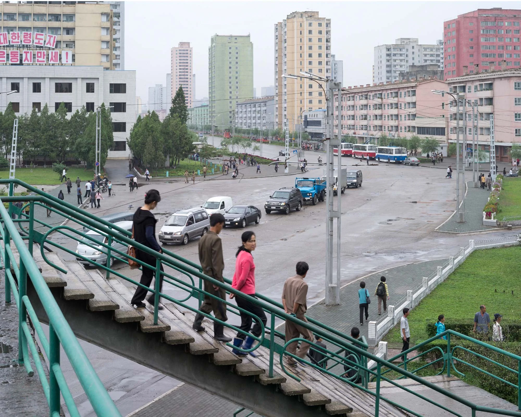 Stadtleben im Zentrum von Pjöngjang, Nordkorea: Passanten überqueren eine Fußgängerbrücke mit grünem Geländer. Im Hintergrund öffnet sich der Blick auf eine breite Hauptstraße mit Autos, Oberleitungsbussen und den für die nordkoreanische Hauptstadt t