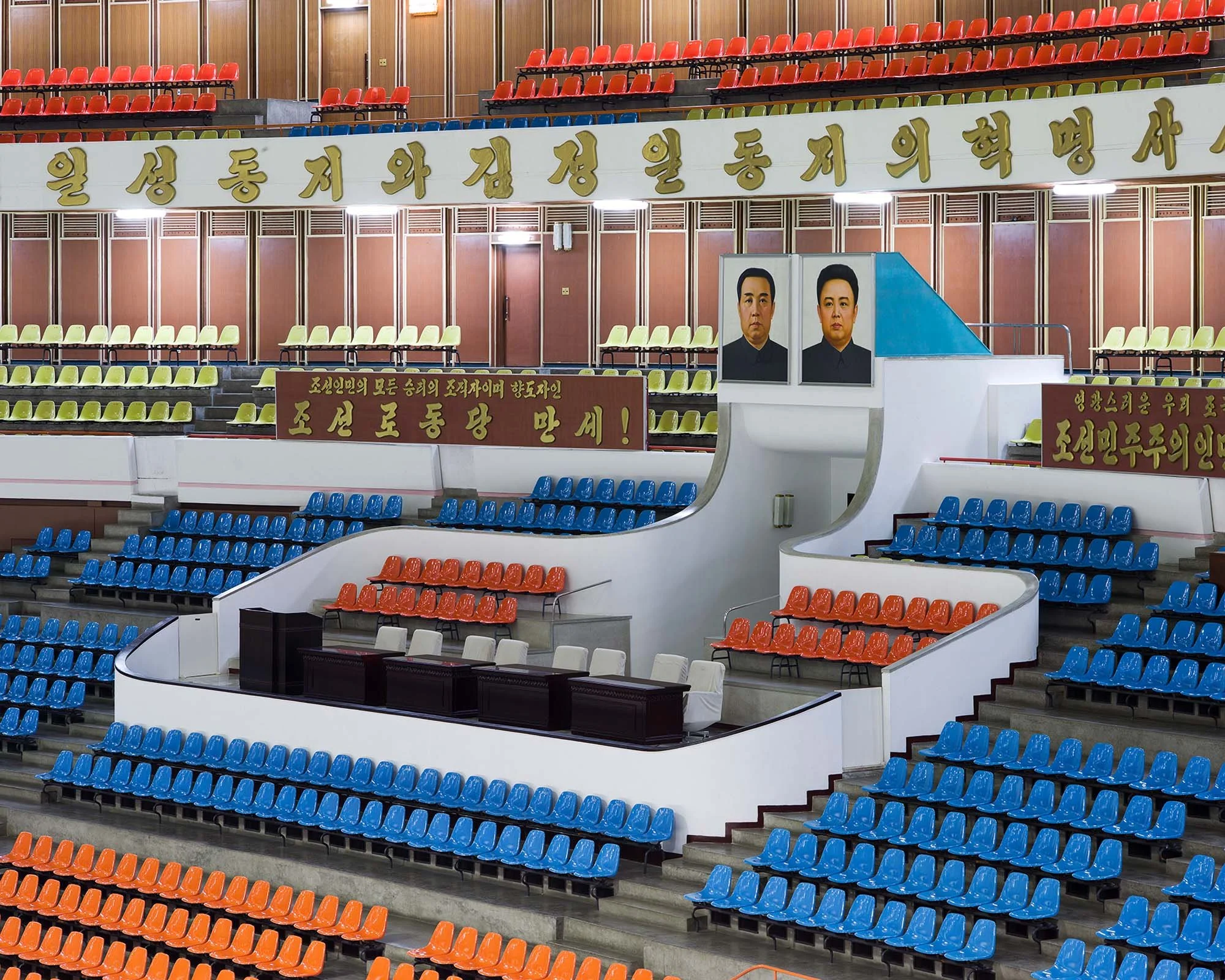 Innenansicht der Eissporthalle in Pjöngjang, Nordkorea: Die markante Arena, die von außen durch ihre kegelförmige Architektur besticht, bietet eine riesige Tribünenlandschaft in Blau und Rot. Im Zentrum steht eine Ehrentribüne vor großen Porträts der