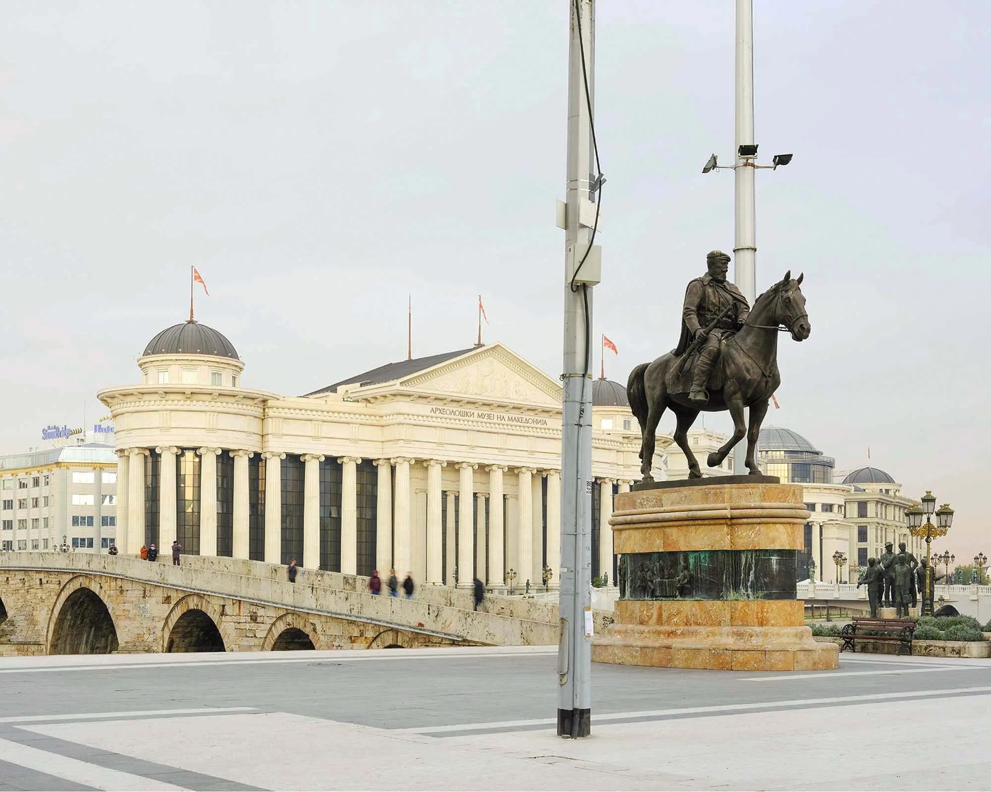 Platz mit Statue eines Reiters vor einem neoklassischen Gebäude mit Kuppeln und Flaggen, und Brücke über den Vardar Fluss  im Vordergrund, an einem bewölkten Tag. Rekonstruktionen in Skopje im Rahmen des Bau- Projektes  Skopje 2014.