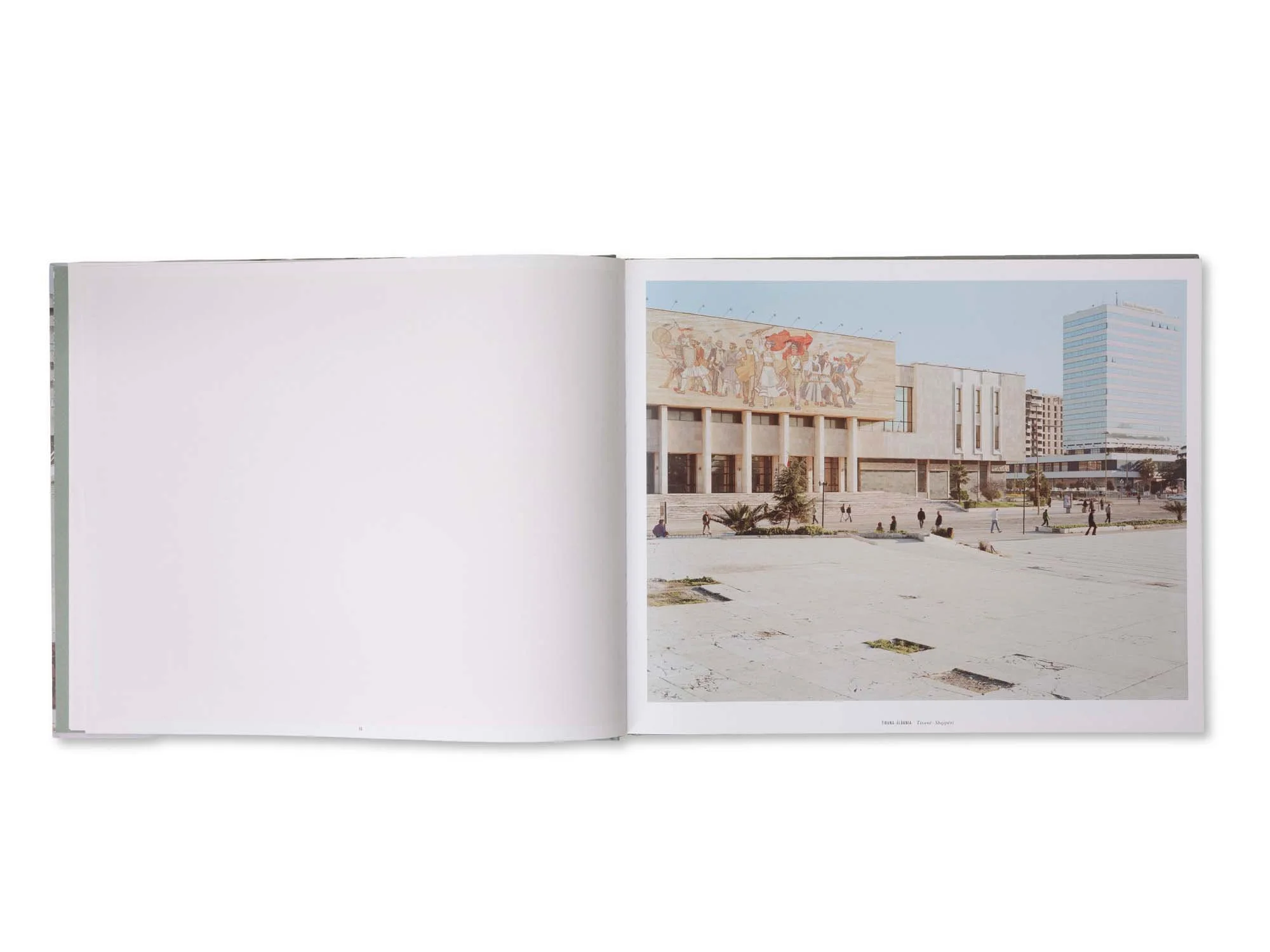 Ein offenes Buch mit einer Fotografie, die eine große Verwaltungsgebäude-Komplex in Tirana, Albanien zeigt. Das Gebäude hat ein Wandgemälde mit tanzenden Figuren an der Fassade. Im Vordergrund sind Menschen auf dem Platz zu sehen.