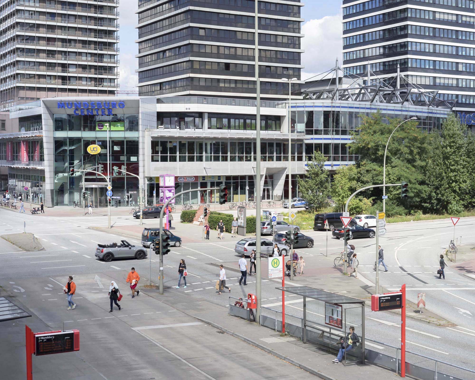 U-Bahn-Station U MUNDSBURG mit mehreren Menschen, die den Bahnsteig betreten und verlassen, umgeben von Hochhausgebäuden, Bäumen und Verkehrsfahrzeugen.