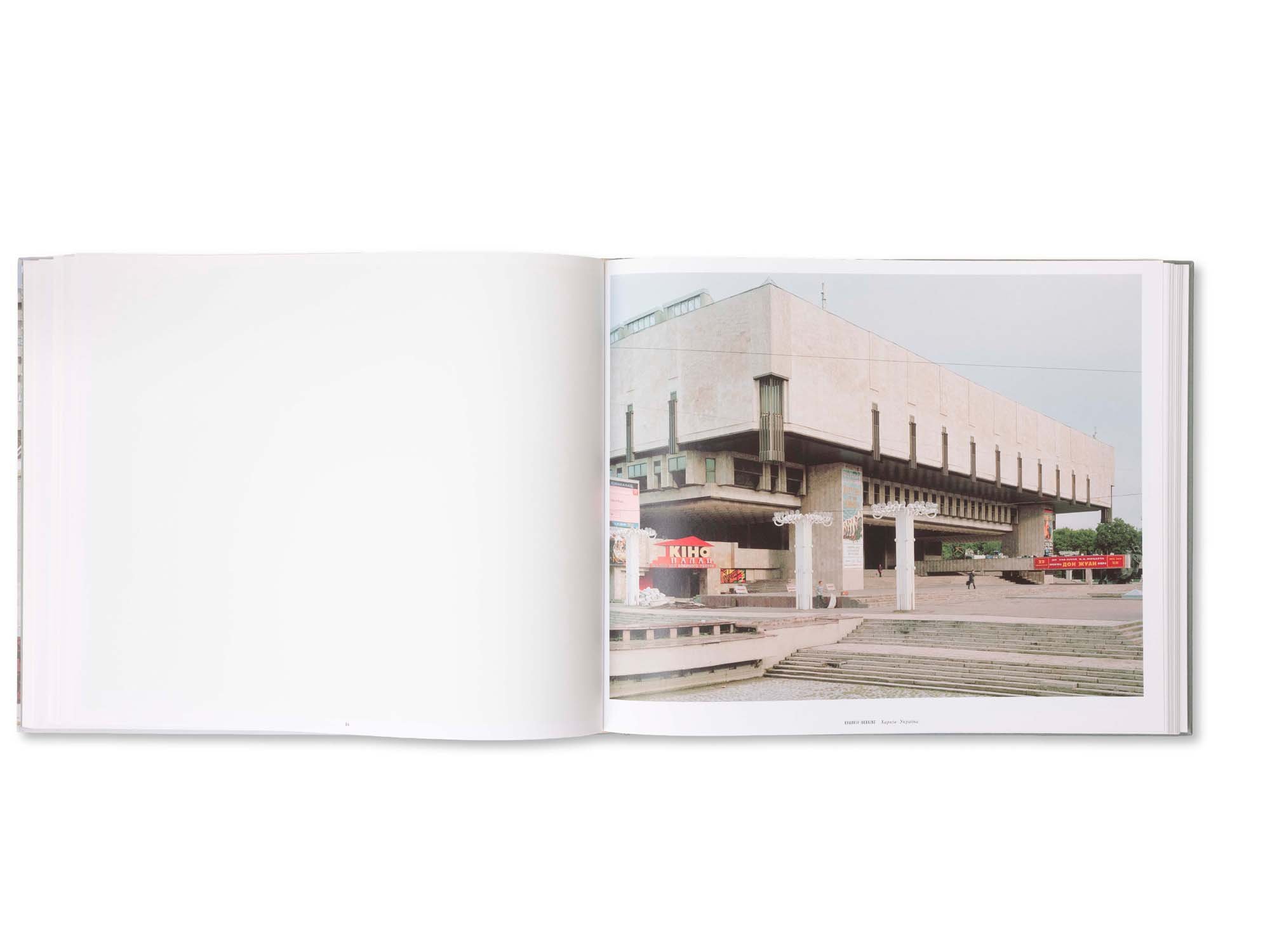 Ein offenes Buch mit einer Fotografie eines modernen Gebäudes auf der rechten Seite. Das Gebäude hat eine kantige, geometrische Architektur mit mehreren Ebenen und Stützen, und befindet sich in einem urbanen Umfeld mit Bänken und Straßenreklamen im V