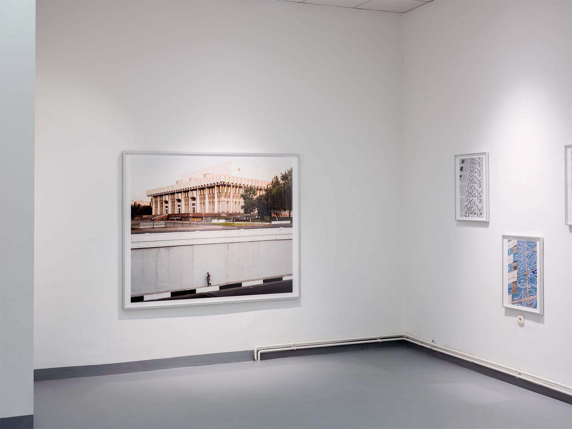 Blick in eine Kunstgalerie mit weißen Wänden, auf denen Fotografien und Kunstwerke hängen. Es sind mehrere Bilder, darunter ein großes Bild eines Gebäudes, das auf der linken Seite hängt, und einige kleinere Bilder rechts. Der Raum ist schlicht und m