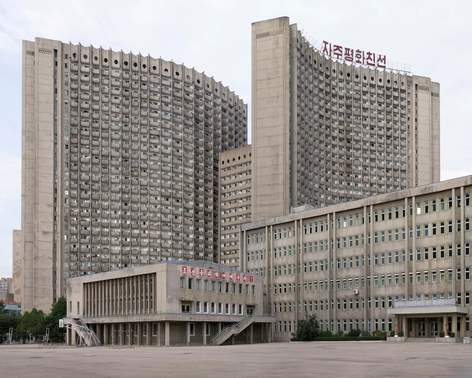 Schulgebäude und Wohnkomplex im Stadtzentrum von Pjöngjang, Nordkorea : Blick über den Vorplatz einer zentralen Schule auf die dahinterliegenden, geschwungenen Wohnhochhäuser im zentralen Bezirk Chung-guyok. Die Dachaufschrift des Brutalismus-Komplex