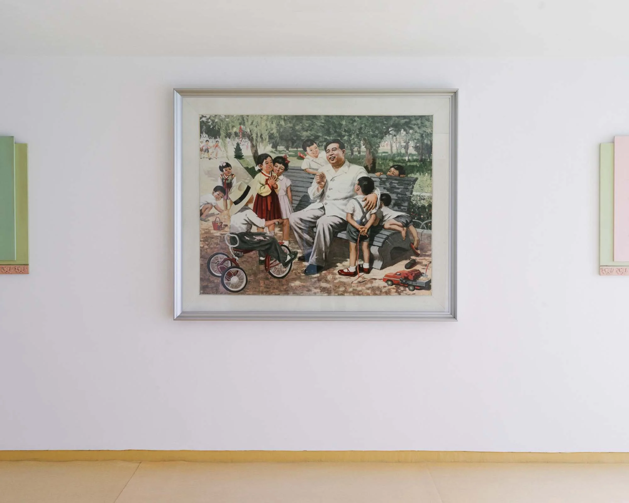 Gemälde von Kim Il-sung in einem Kindergarten in Pjöngjang, Nordkorea: Ein großformatiges Propagandagemälde an einer minimalistischen Wand zeigt den Staatsgründer Kim Il-sung von lachenden Kindern und Spielzeug umgeben auf einer Parkbank. Solche idea