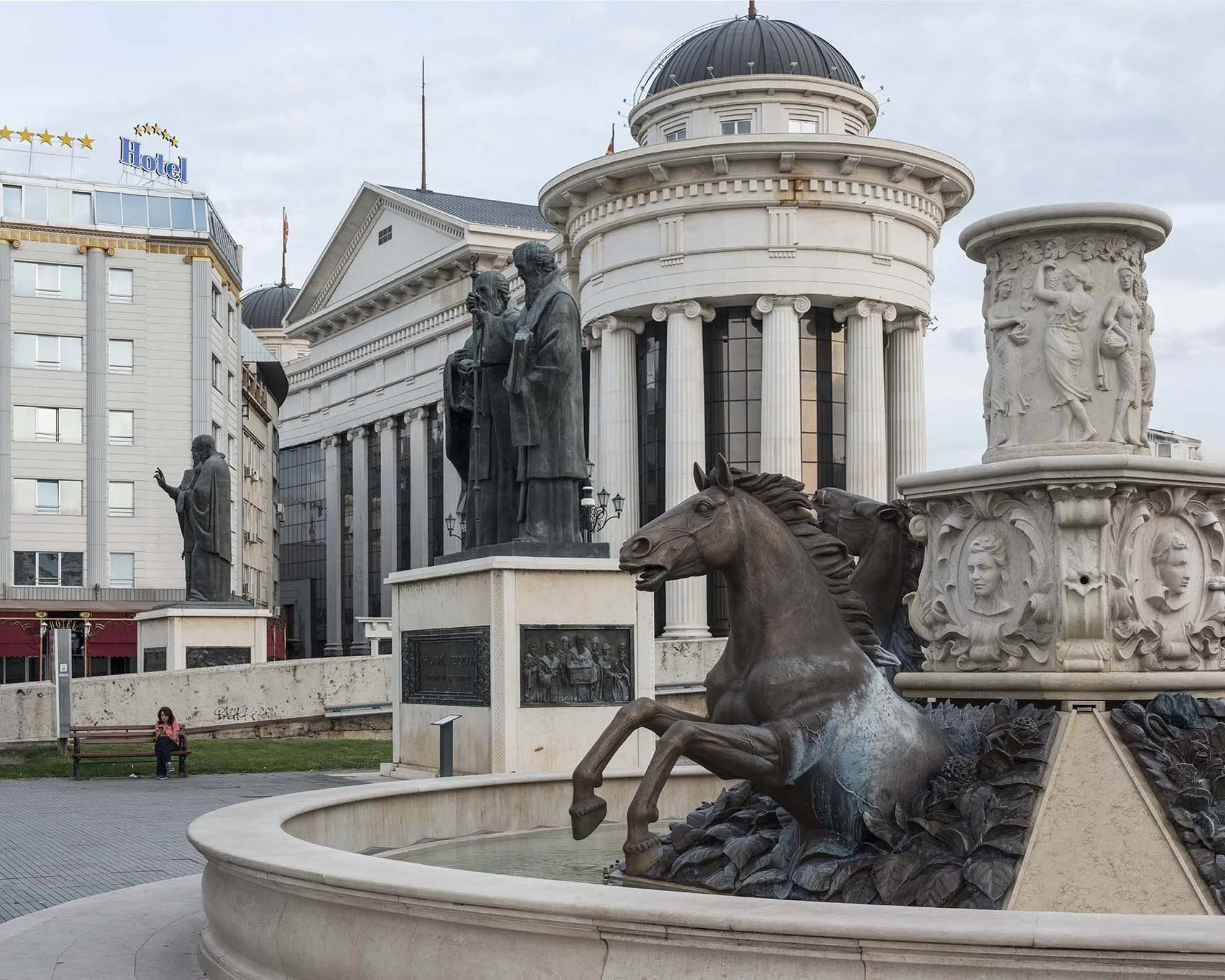 Statuen und Skulpturen in einem öffentlichen Platz, umgeben von Gebäuden, darunter ein Hotel. Die Skulpturen zeigen historische Figuren und ein Pferd.