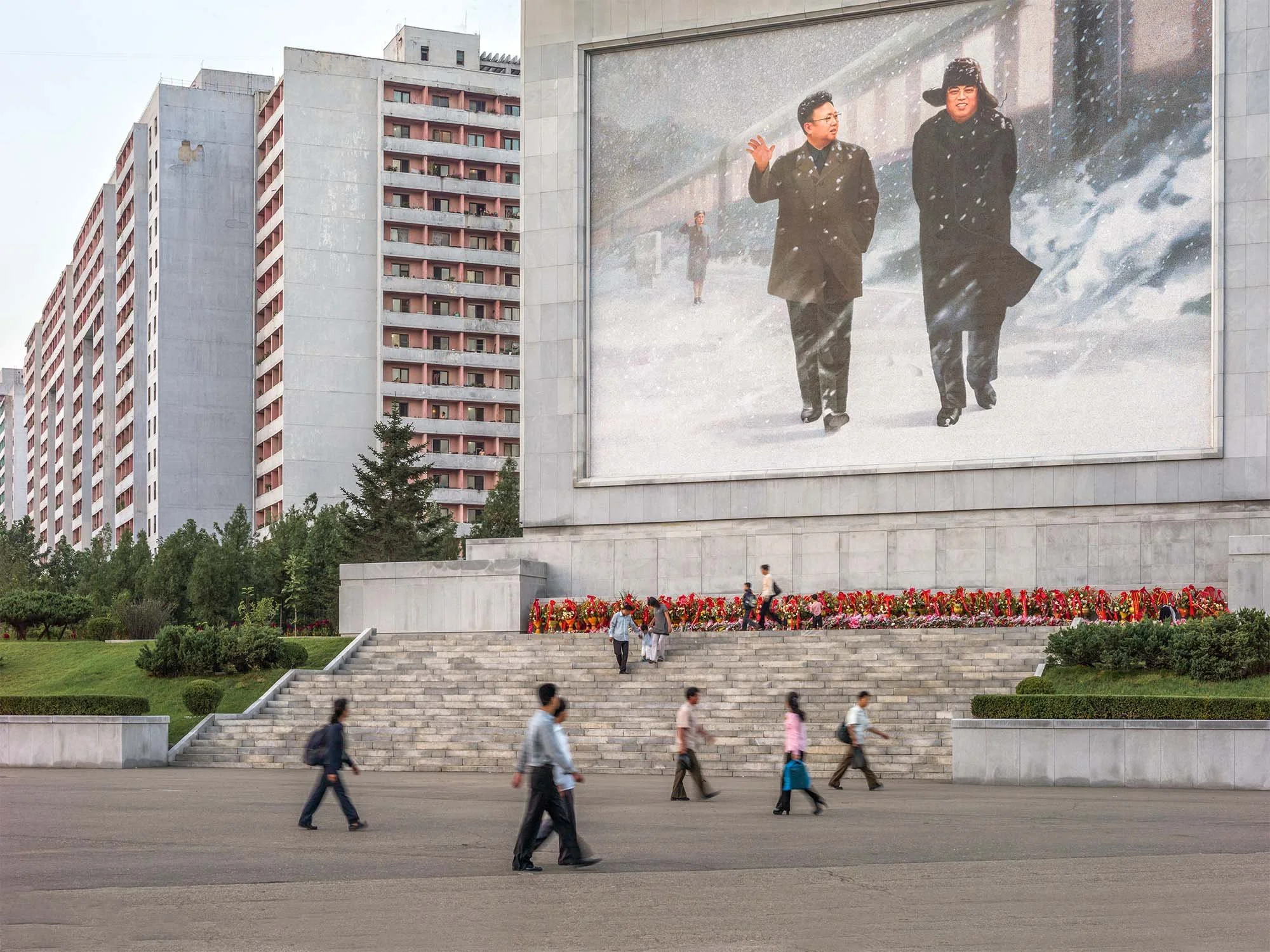 Wohnquartier in Pjöngjang mit Monumentalwandbild, Nordkorea: Im Vordergrund des Wohnkomplexes steht eine gigantische Wandtafel mit einer Mosaikdarstellung von Kim Il-sung und Kim Jong-il im Schnee. Passanten kreuzen das Areal vor den typischen pastel