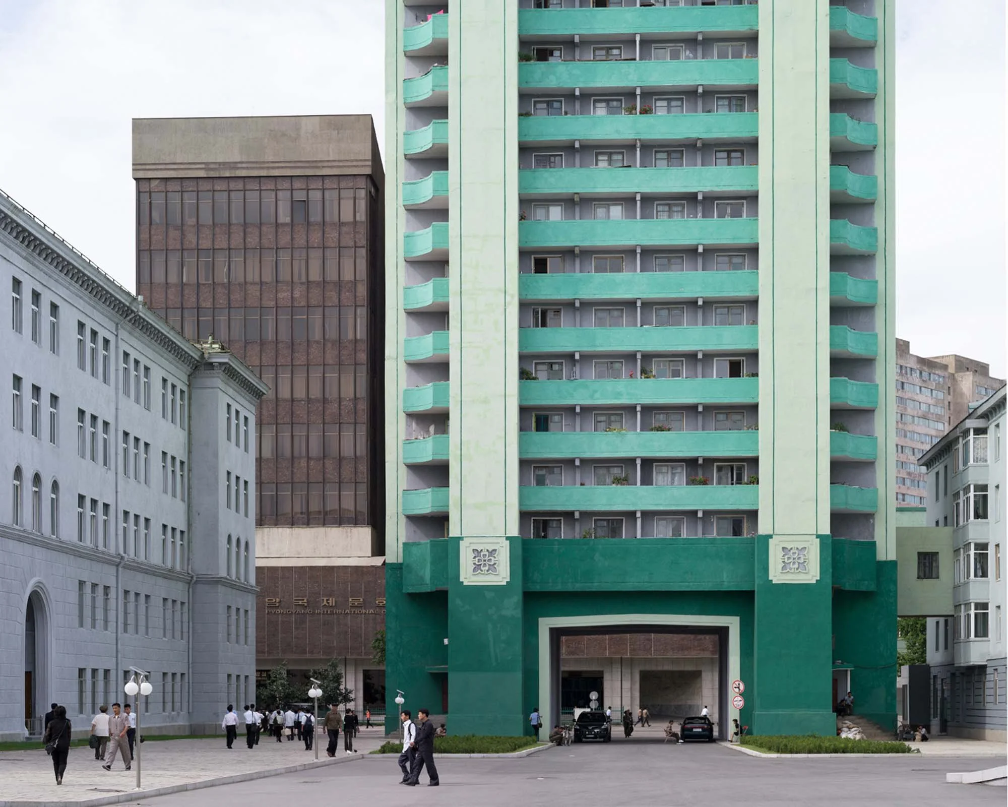 Moderne Stadtarchitektur im Zentrum von Pjöngjang, Nordkorea: Ein auffälliges mintgrünes Wohnhochhaus mit großer Tordurchfahrt prägt das Straßenbild nahe dem Pyongang International Communications Center (im Hintergrund). Passanten flanieren über den 