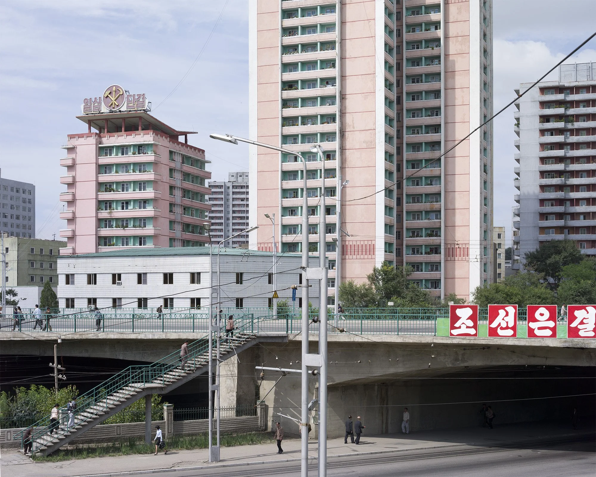 Stadtarchitektur im Zentrum von Pjöngjang, Nordkorea: Der Blick zeigt eine Überführung mit Treppenaufgang sowie charakteristische rosafarbene Hochhäuser. Das linke Gebäude wird von dem Emblem der Partei der Arbeit Koreas (Hammer, Sichel und Pinsel) g