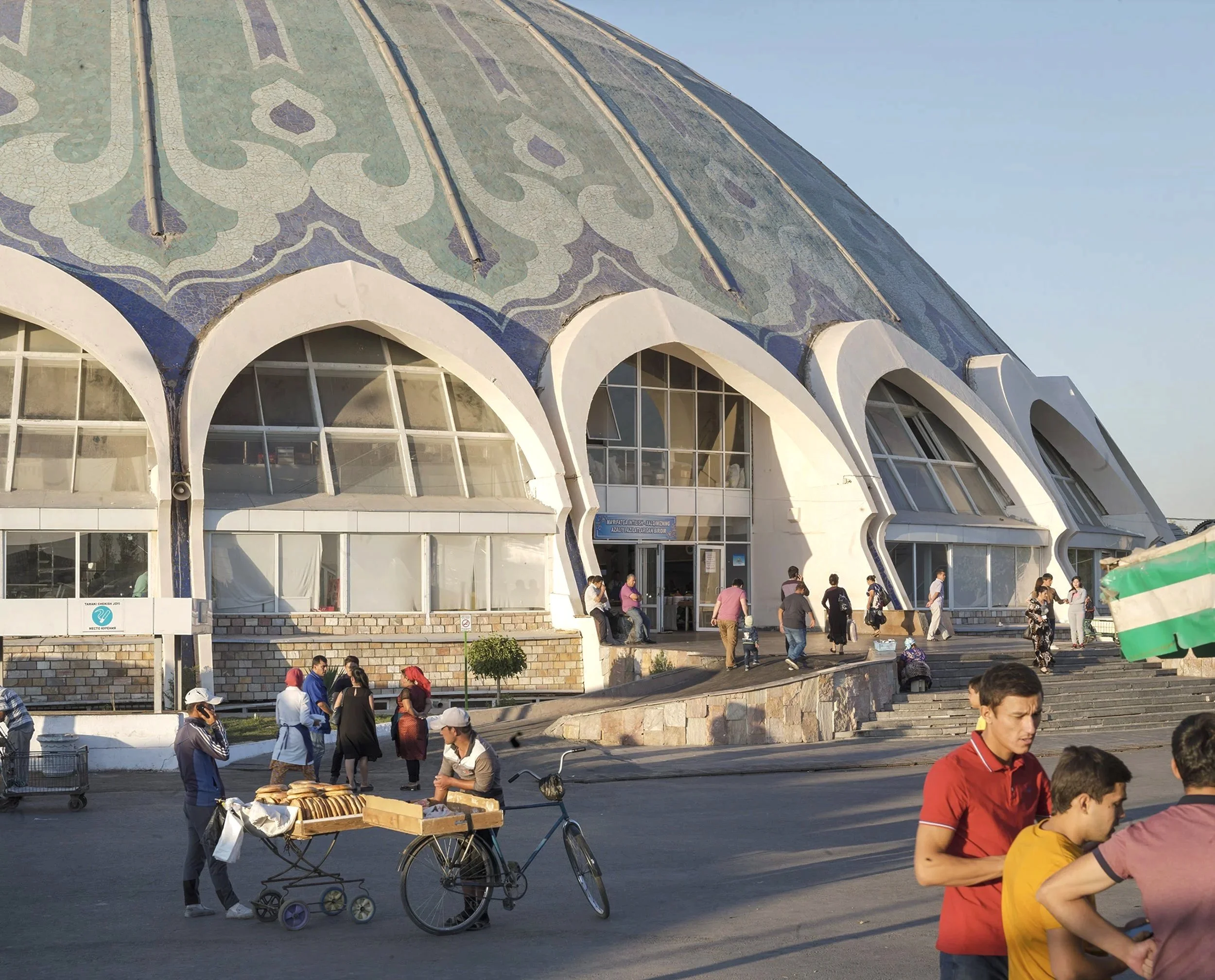 Menschen versammeln sich vor einem modernen Gebäude mit einer wellenförmigen Fassade und Mosaic-Mustern auf dem Dach, sichtbar sind Verkäufer mit Brot und Passanten, einige stehen oder gehen.