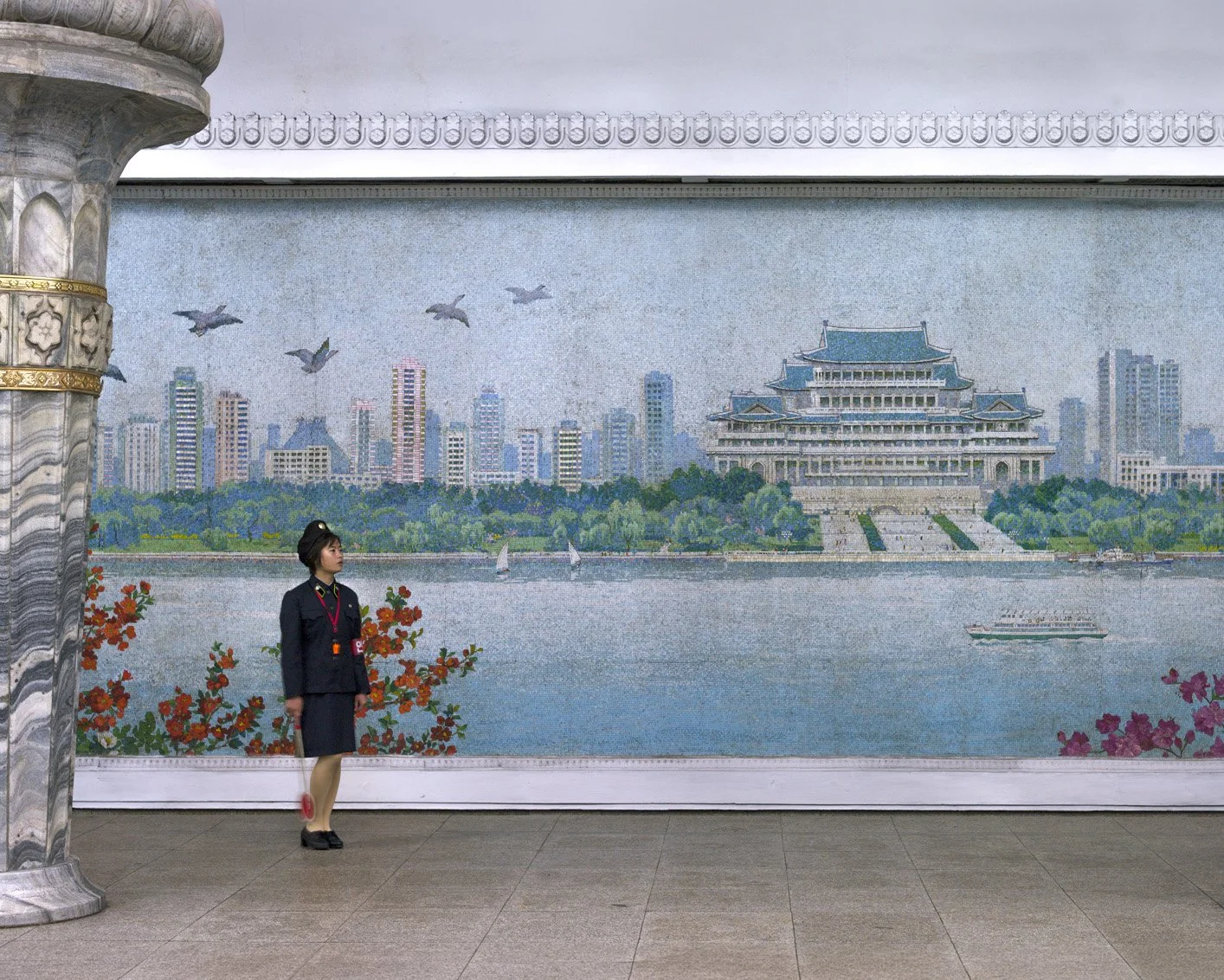 U-Bahn-Station der Metro Pjöngjang in Nordkorea: Eine Bahnsteigschaffnerin in Uniform steht mit Signalkelle vor einem imposanten, wandfüllenden Mosaik. Das Kunstwerk zeigt eine städtische Panoramaansicht mit dem Großen Palast der Volksstudien am Ufer