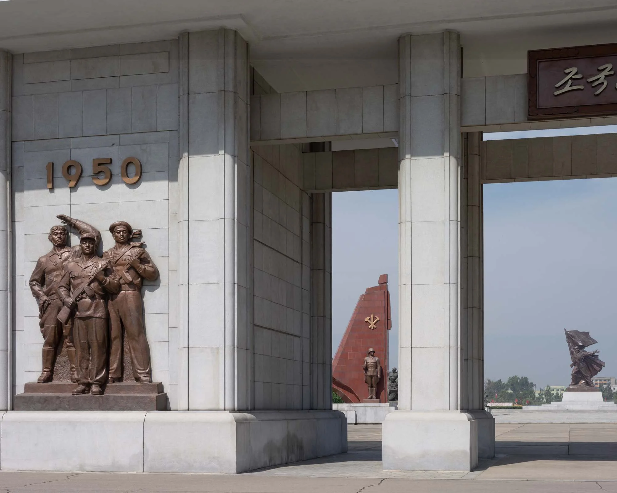 Museum über den Vaterländischen Befreiungskrieg in Pjöngjang, Nordkorea: Der Außenbereich des Kriegsmuseums zeigt bronzene Reliefs von Soldaten unter der Jahreszahl 1950, dem Beginn des Koreakriegs. Durch die Säulenkonstruktion des Monuments blickt m